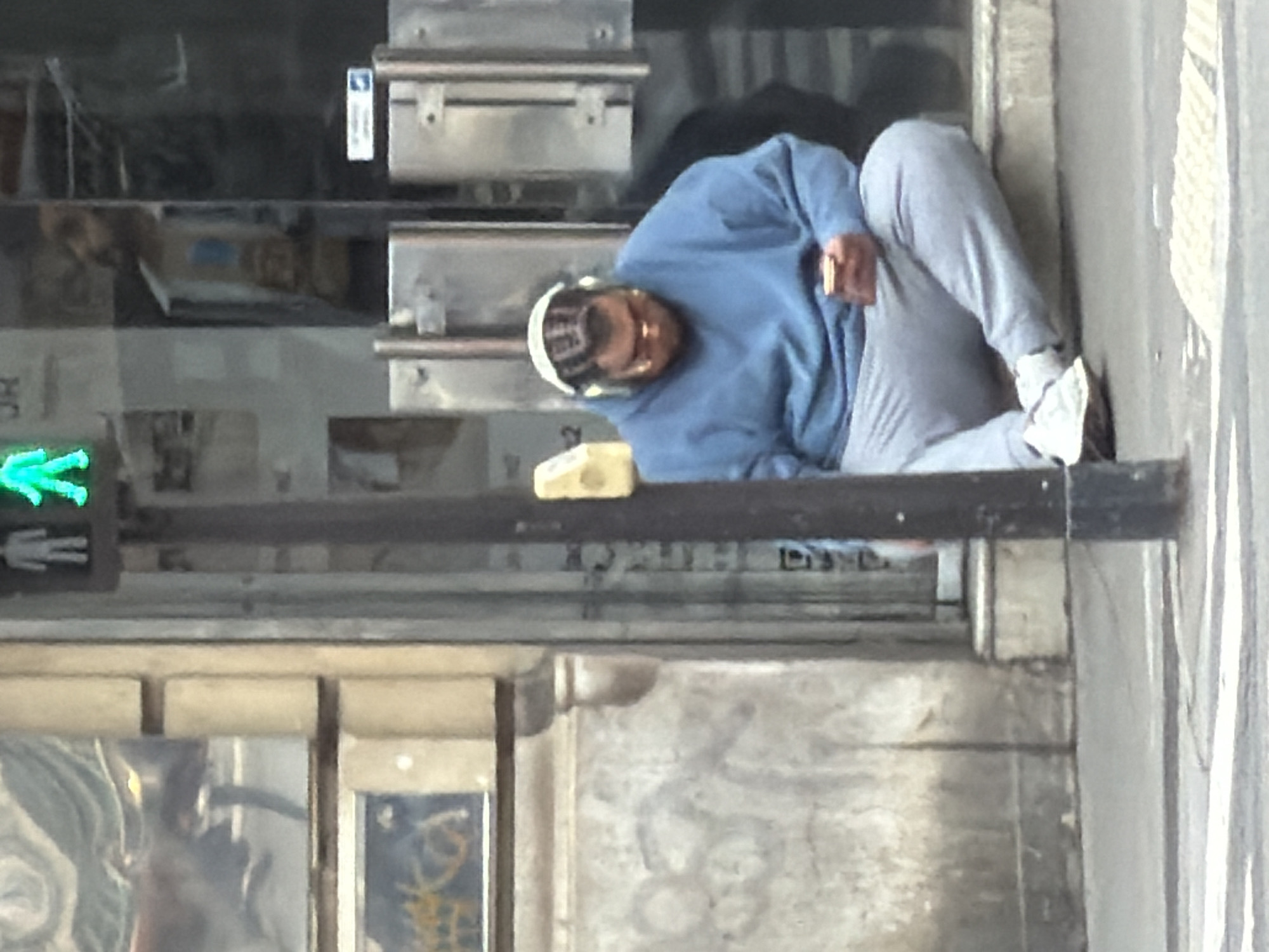 A medium, rotated, outdoor shot depicts a person reclining on an elevated ledge, apparently in Paris, France. The individual is light-skinned, clad in a light blue sweatshirt and light grey sweatpants, with white footwear. The person is sitting and holding an object in their right hand. They wear a white hat or head covering. The ledge they are sitting on runs horizontally through the frame, with a traffic signal visible to the left. Behind the ledge is a metal structure, possibly related to a storefront. The architecture visible includes signs of urban wear and age. The street and a portion of the sidewalk are to the right of the frame.