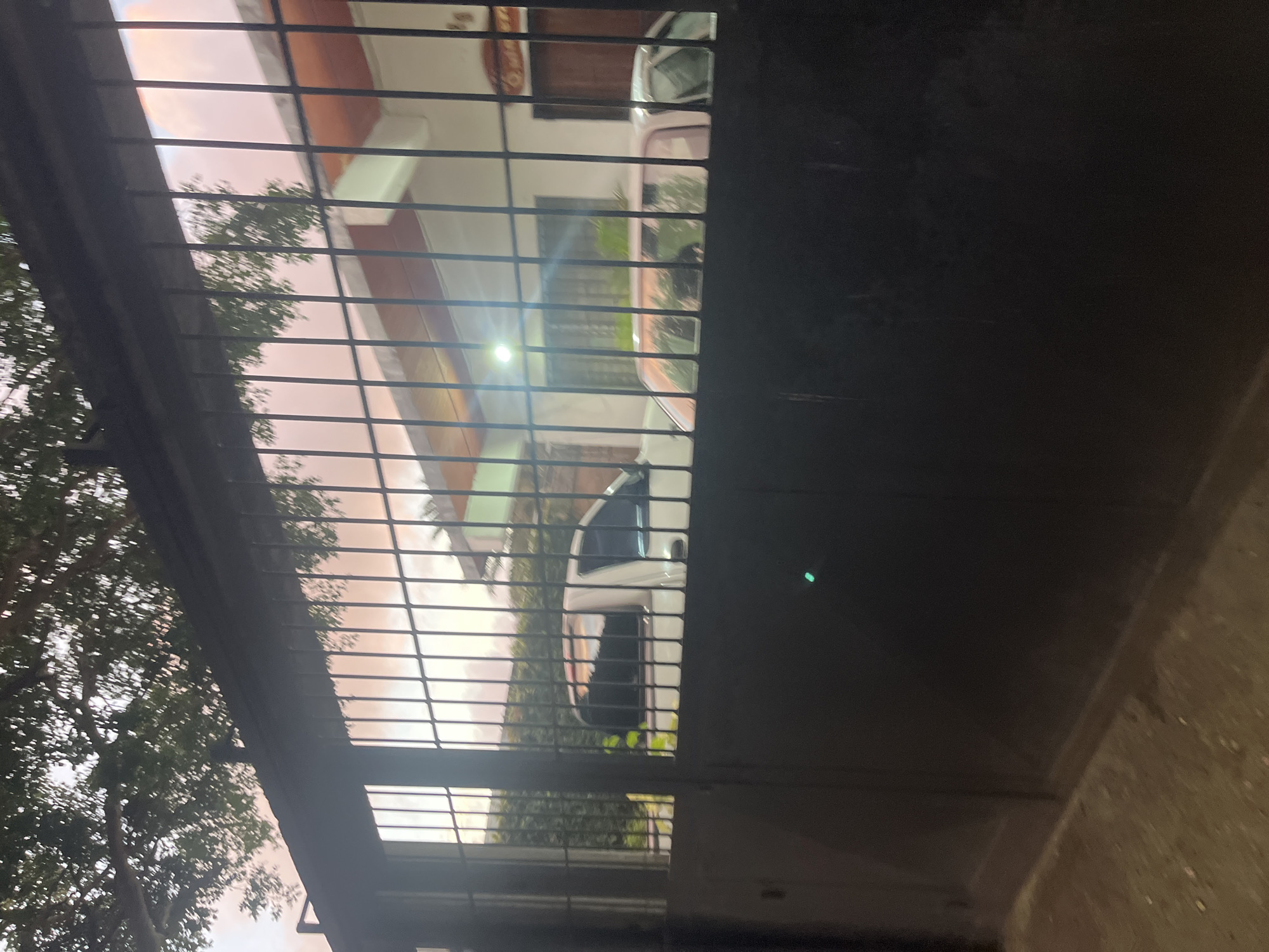 The image shows a view through a black metal gate at a residential property in Santa Ana, Venezuela. A white pickup truck is parked on the left side of the driveway, and a light-colored car is parked further back on the right. The house is visible in the background, with a light on inside. The sky appears to be a pale pink and orange, suggesting it is either dawn or dusk. There are trees and foliage surrounding the property. The foreground shows the dark metal of the gate and a dusty ground surface.