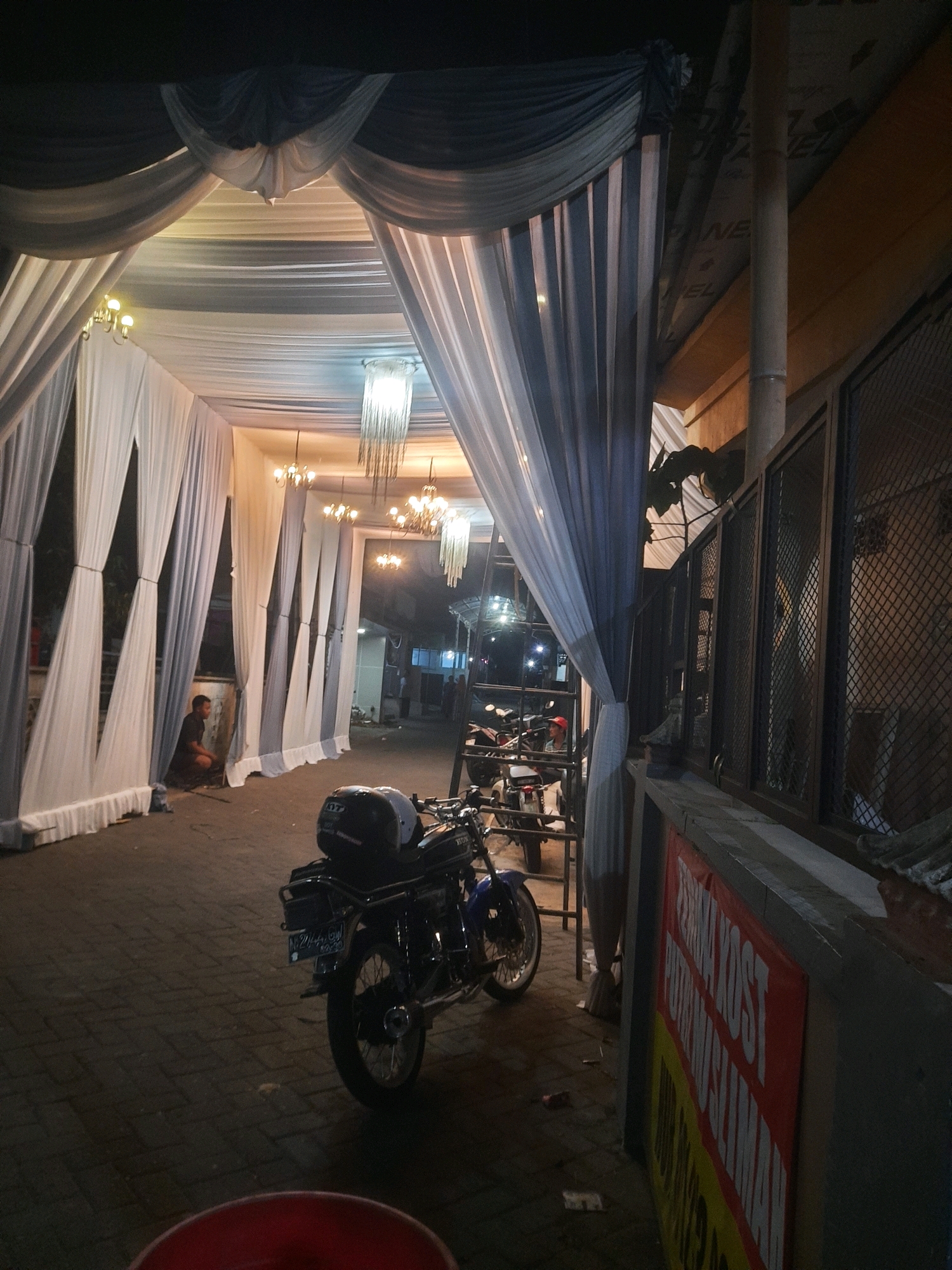 This is a night-time, eye-level, outdoor photograph taken in the city of Kepuharjo, Indonesia. The scene is dominated by a draped tent or canopy structure, likely for an event, with white and grey fabric. Several decorative chandeliers hang from the ceiling of the canopy. In the foreground, a black motorcycle is parked on a paved walkway, with a black and white helmet resting on the seat. A person is partially visible, squatting in the background to the left, near a wall covered by white fabric. To the right of the motorcycle, a metal fence with diamond-shaped openings is visible, partially obscuring structures behind it. A red sign with white and yellow text is mounted on a wall in the lower right corner of the image. The ground appears to be paved with interlocking bricks. The lighting suggests artificial light sources within the canopy and possibly from surrounding buildings.