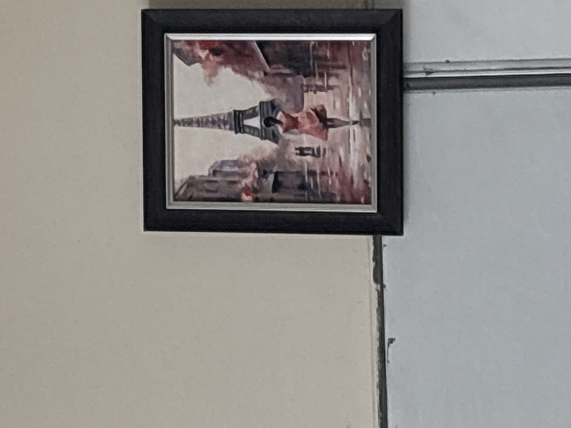 A framed picture hangs on a wall. The picture depicts an artistic rendering of the Eiffel Tower and figures, possibly in Paris. The frame is black. The wall behind it appears to be light-colored, possibly cream or white. A metal rod is visible in the upper right of the image.