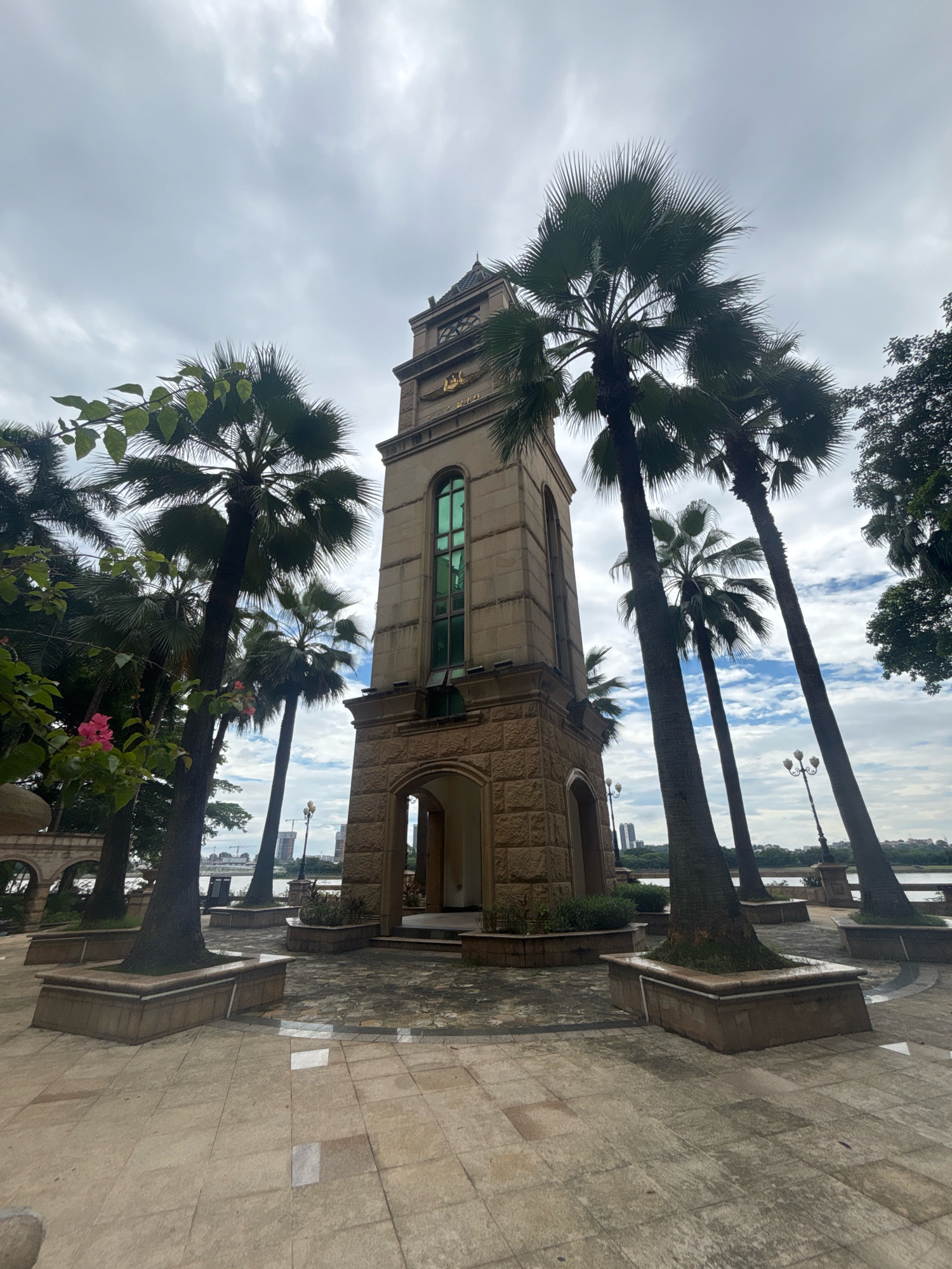The image depicts a stone clock tower with a green window and decorative golden emblems, situated in a plaza with palm trees and patterned paving stones. The clock tower is the central subject, with its weathered stone facade and arched entrance. Several tall palm trees are arranged around the plaza, some framing the clock tower. In the background, a body of water is visible, beyond which lies a cityscape with buildings and cranes under a cloudy sky. The paving stones in the foreground are light-colored and appear wet in places, reflecting the overhead light. Decorative planters with greenery are interspersed within the plaza. The scene is devoid of people. The location is identified as the clock tower in Nanhai District, China.