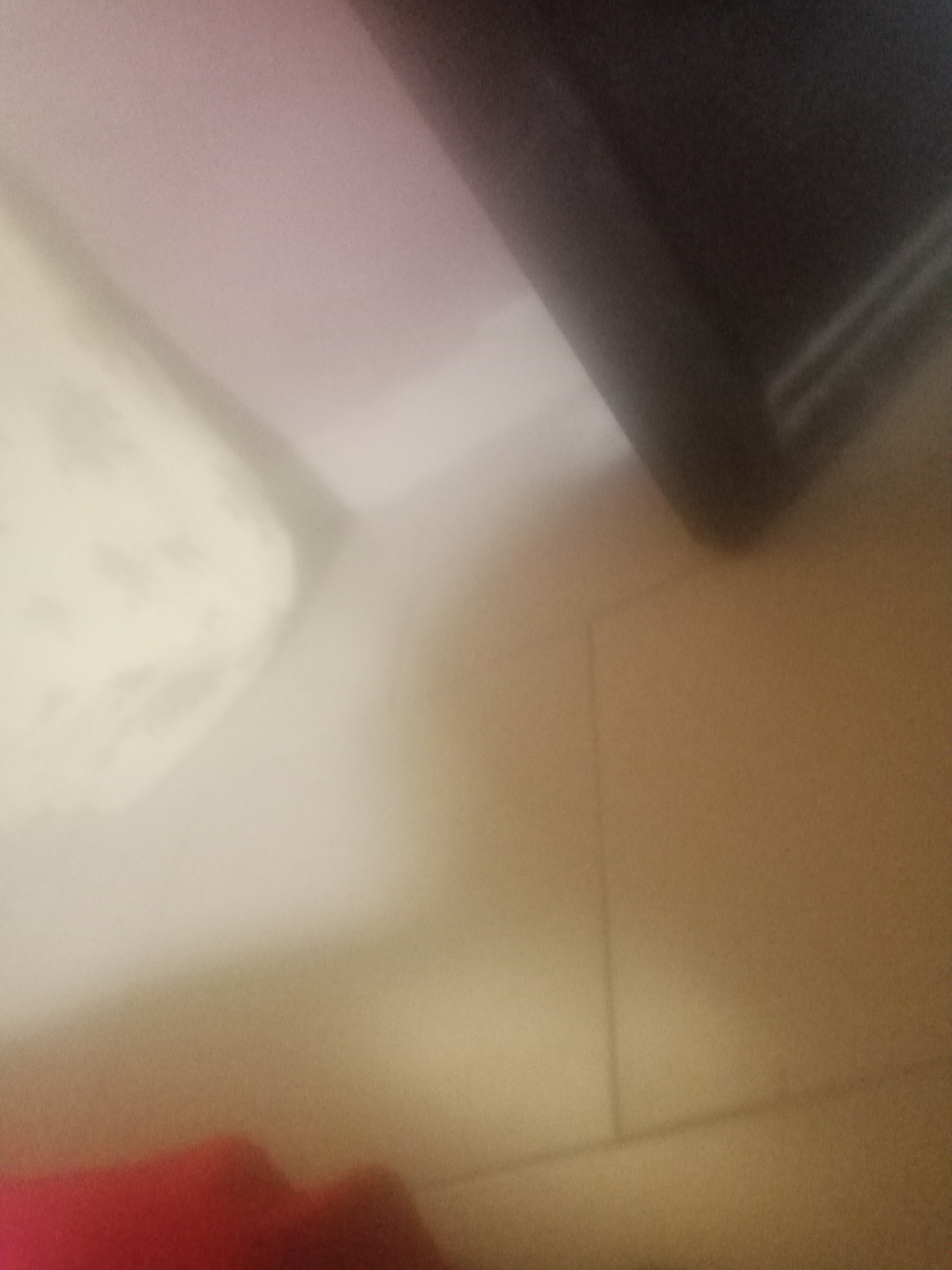 The image shows a dimly lit, close-up view of a surface that appears to be a floor or table. The dominant colors are muted tones of brown, beige, and a soft pink. In the upper left, there is a section of a light-colored fabric with a subtle pattern, possibly a blanket or clothing. Adjacent to this, and extending towards the top right, is a dark, solid object, possibly a piece of furniture or an electronic device. The lighting creates soft shadows and gradients across the surfaces. In the bottom left corner, a patch of vibrant red is visible, suggesting another fabric item. The overall impression is of an indoor setting with diffuse lighting.