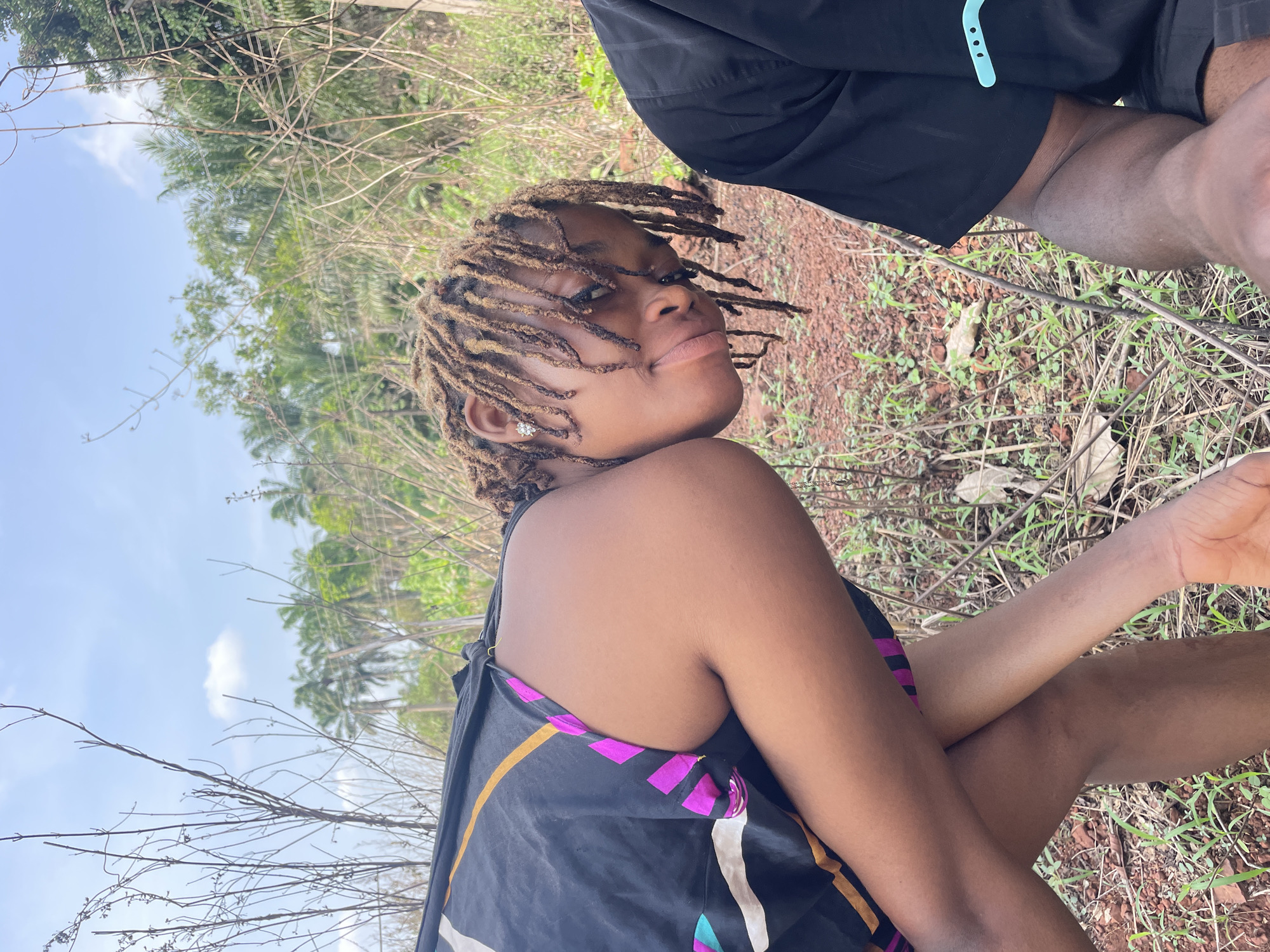 A young woman with light brown dreadlocks is the primary subject of the image, looking back over her left shoulder towards the camera with a slight smile or smirk. She is positioned towards the left side of the frame, appearing to be sitting or crouching with her arms crossed, revealing a black sleeveless top adorned with vibrant geometric patterns in pink, yellow, white, and teal. A small, sparkly earring is visible in her left ear.
The setting is outdoors, characterized by reddish-brown soil, sparse green ground cover, and numerous dry, bare branches and twigs scattered in the foreground and midground. In the background, lush green foliage and palm trees indicate a tropical or semi-tropical environment. The sky above is clear blue with a few faint white clouds, suggesting a bright daytime setting.
Part of another person is visible in the upper right portion of the image, wearing black shorts. A light blue strap or accessory is attached to their clothing near the thigh. The overall impression is a candid outdoor shot, possibly a moment of relaxation or a casual photoshoot in a natural, possibly rural, area consistent with the location context of Ogbe, Nigeria. There is no visible text in the image.