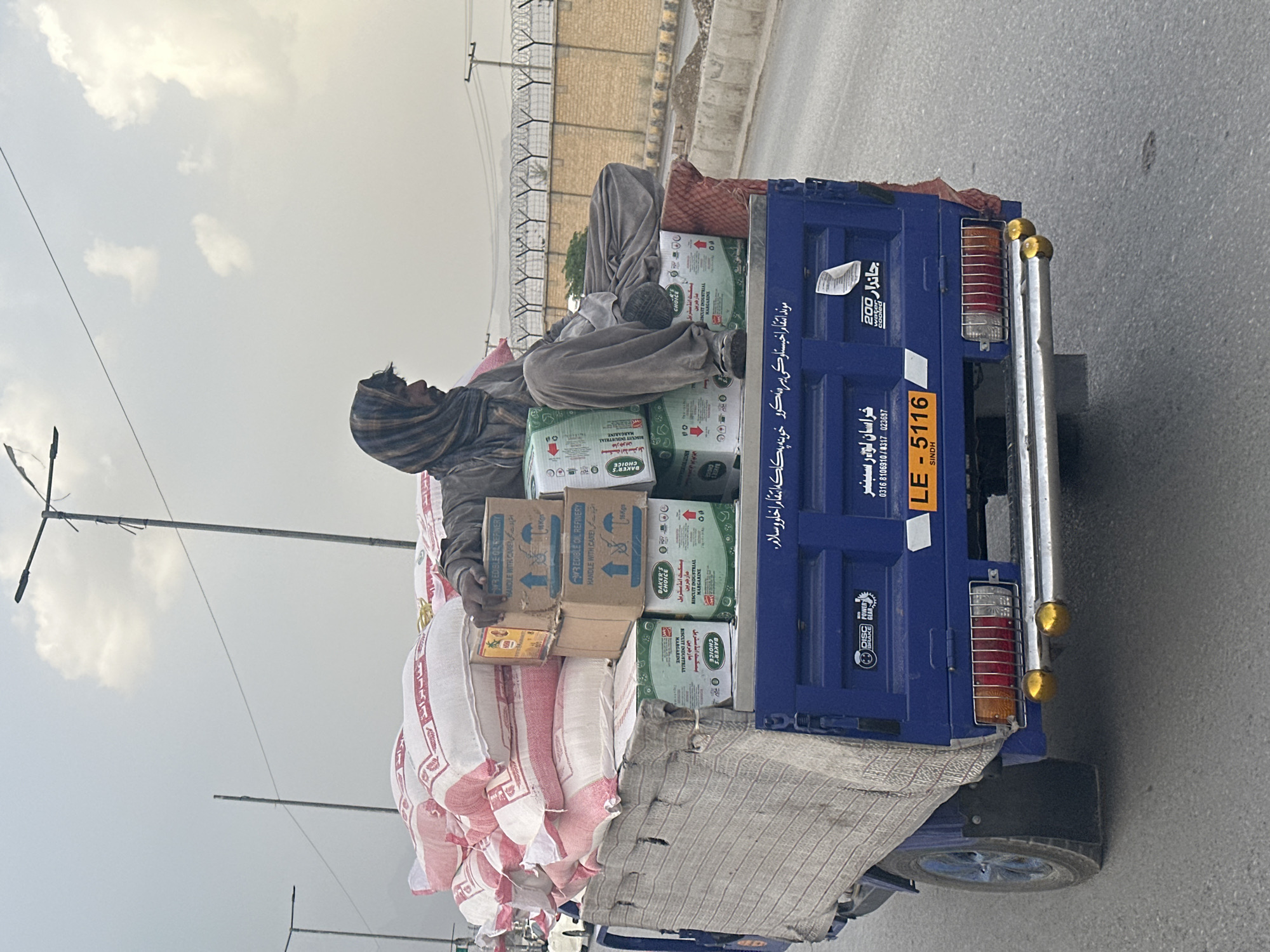 A low-angle, eye-level shot shows a man riding atop a heavily laden vehicle, potentially a small truck or trailer, on a street in Quetta City Tehsil, Pakistan. The man is partially covered with cloth and sits atop numerous sacks, likely filled with goods. Above the sacks are stacked boxes labeled "Baker's Choice Biscuit Industrial Margarine" and "Refined Edible Oil Refinery."
The vehicle is dark blue with tail lights visible, and the license plate reads "LE-5116 Sindh." Writing in a non-English script is visible on the side of the vehicle. The surrounding environment consists of an urban landscape with a road, light poles, and a building with barbed wire fencing in the background. The sky is overcast. The man appears to be resting.