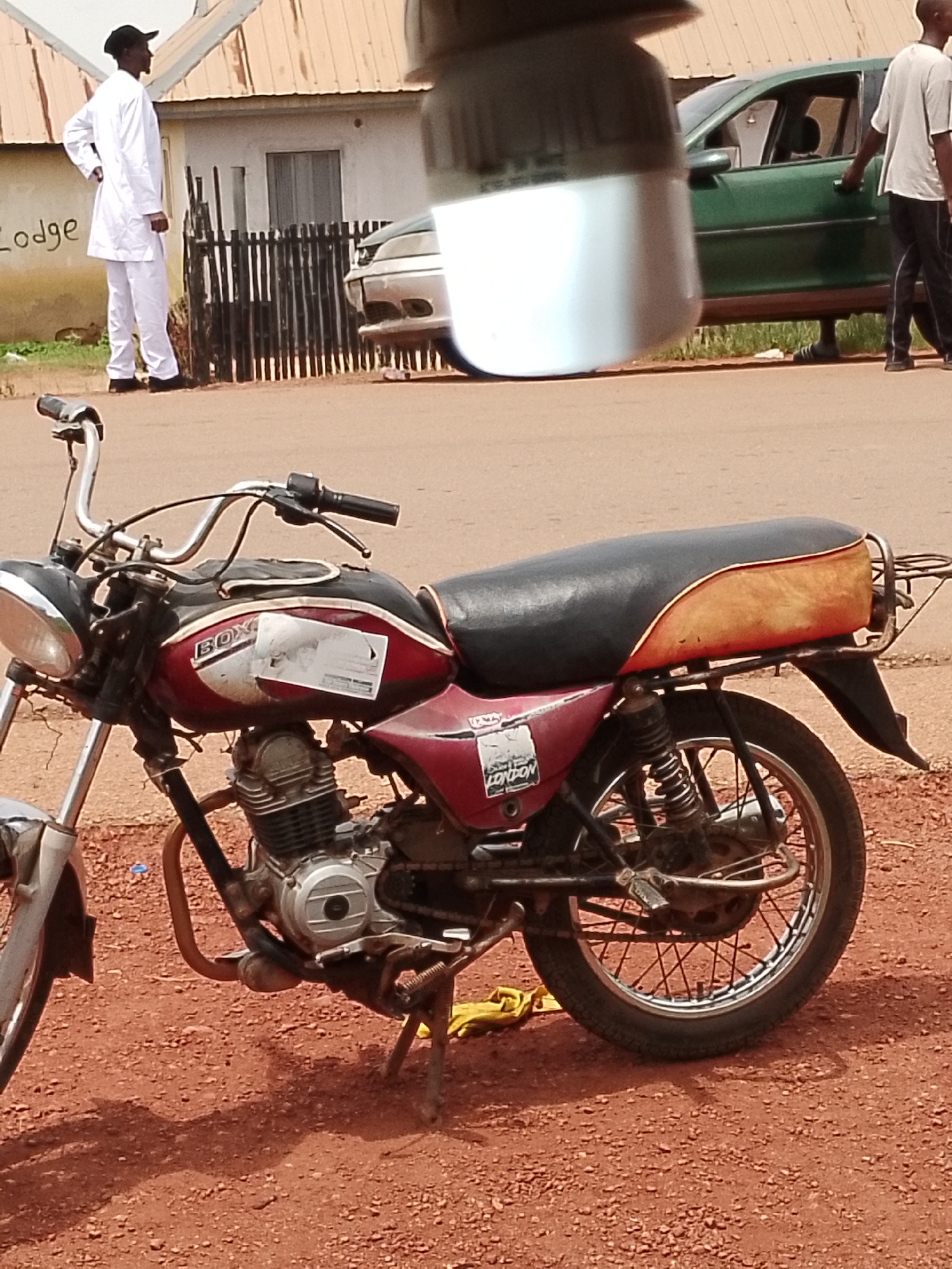 In Wumat, Nigeria, a red and black motorcycle is parked on a dirt road in front of a building. A man in a white tunic and pants, wearing a black cap, stands to the left of the motorcycle, facing away from the camera. A dark-colored car is parked behind a dark fence to the right of the building. The building has a light-colored corrugated metal roof and a light-colored wall with the word "LODGE" painted on it. The ground is covered in red dirt and small stones, with shadows cast from the motorcycle and an unknown object in the foreground.