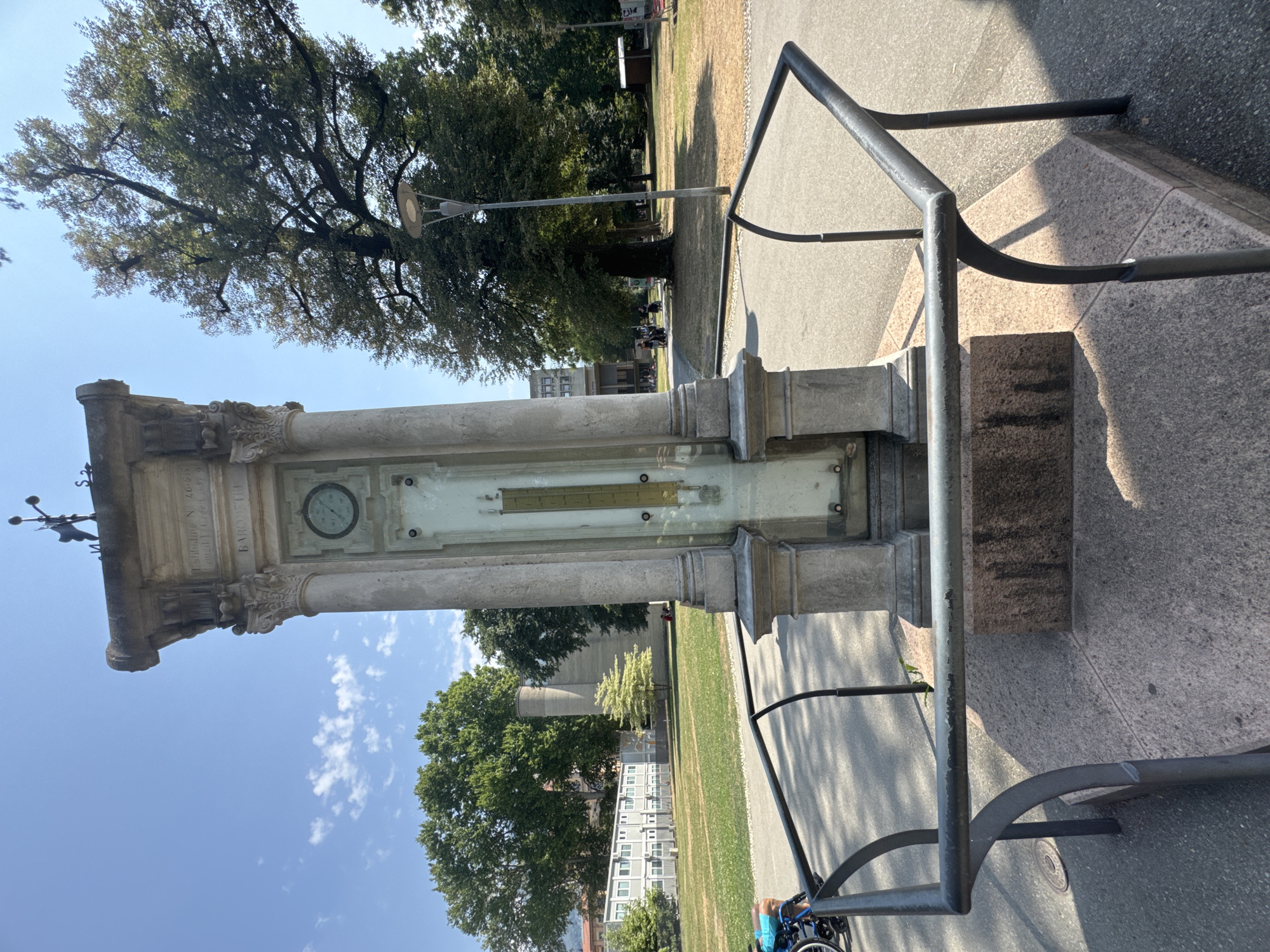 The image shows a weather station located in a park area in Lugano, Switzerland. The weather station is a stone monument with classical architectural elements, including columns and decorative carvings. It features a clock face, a barometer, and a thermometer. In the foreground, there are metal railings surrounding the base of the monument. A person in a wheelchair is visible on a paved path to the left of the monument. In the background, there are trees, buildings, and other park visitors. The sky is blue with some scattered clouds, suggesting a clear and sunny day.