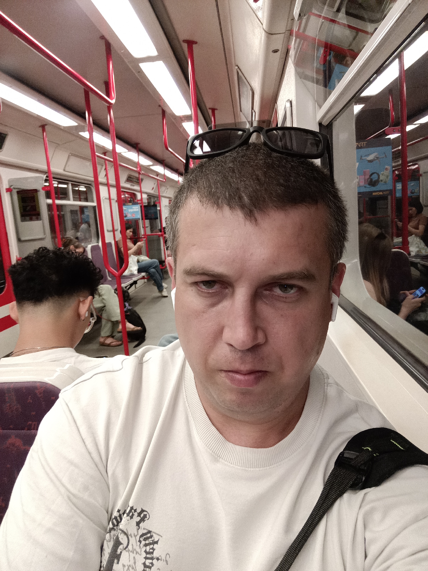 The image is a selfie taken by a man on a Prague metro train. He is wearing a white t-shirt and sunglasses perched on his head. He has an earbud in his right ear and a strap from a bag is visible over his left shoulder. The train carriage is visible in the background, with red poles and fluorescent lighting. Other passengers are seated on purple seats, some looking at their phones. The overall mood is casual, and the time of day appears to be evening, given the artificial lighting. The location is confirmed as Prague, Czechia. There is no visible text in the image.