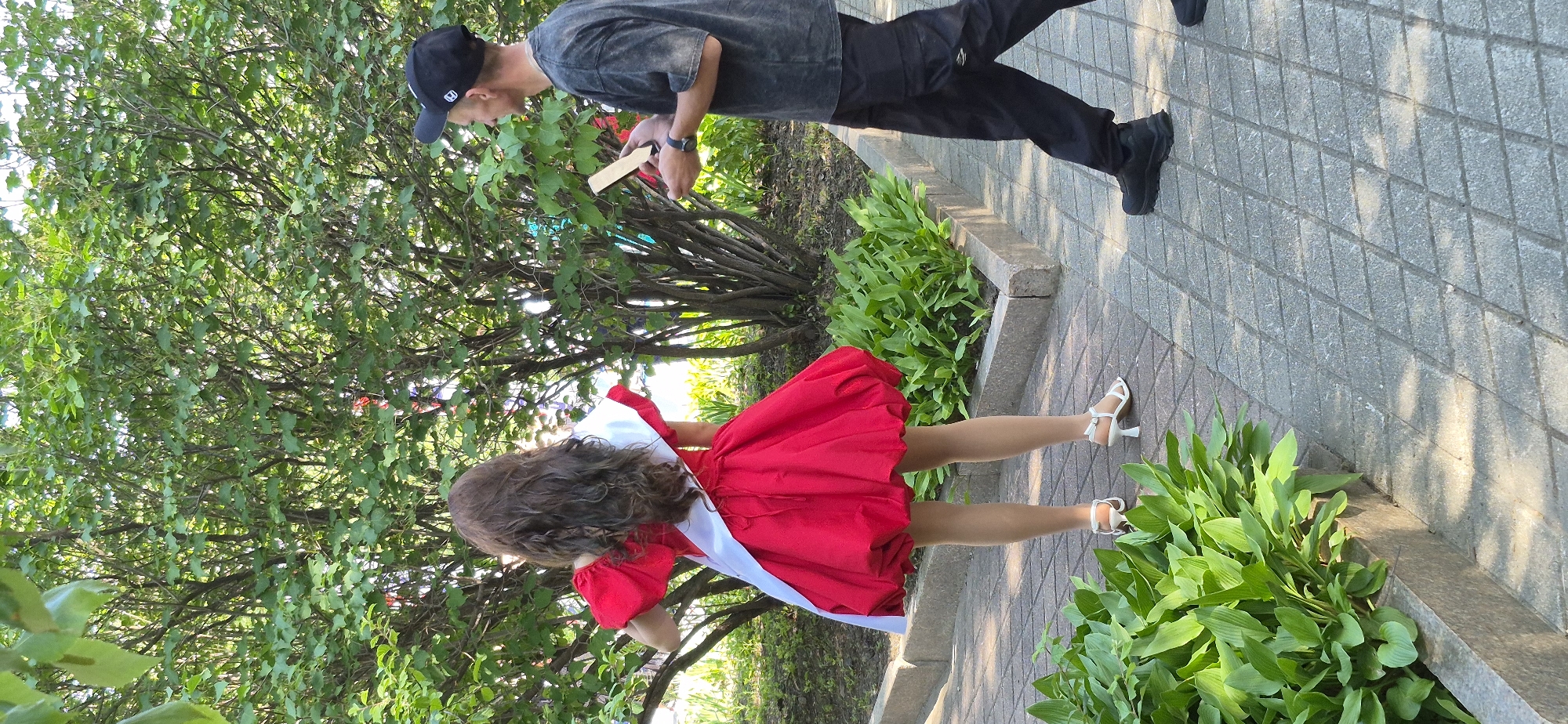 In Ussurijsk, Russia, a person with long, wavy brown hair, wearing a red dress with a white sash and white heeled sandals, stands facing away from the camera, looking towards a leafy tree. To the right, a man in a black baseball cap and dark grey t-shirt holds a rectangular object, possibly a tablet or phone, and appears to be interacting with the first individual. They are standing on a paved walkway with a border of green plants.