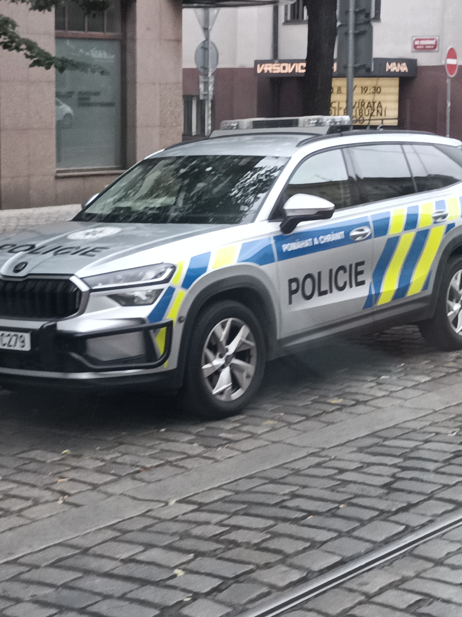 The image shows a silver police car, identified as "POLICIE", parked on a cobblestone street in Prague, Czechia. The car has blue and yellow reflective stripes on its side. In the background, there is a building with a sign that reads "VRSOVIC MANA". Another sign above it displays "8. / 19:30 SLAVIRATA". A traffic light and a red and white circular traffic sign are also visible. The car's license plate is partially visible, showing "CZ09". The scene suggests an urban environment in the Czech capital.