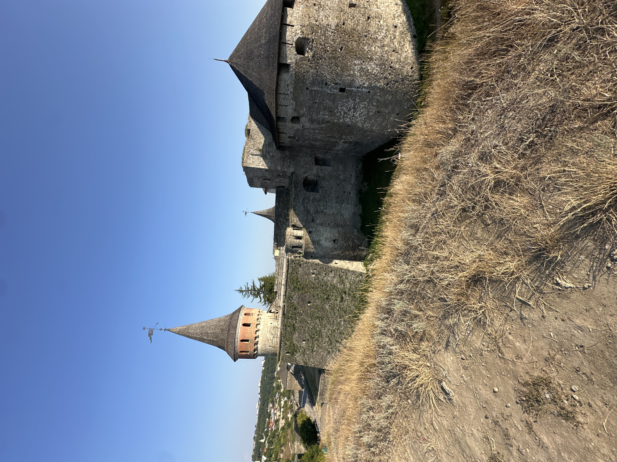 An exterior, eye-level, wide shot captures the Kamianets-Podilskyi Castle in Ukraine under a clear, bright blue sky. The castle, constructed from rough-hewn stone, features thick, imposing walls and several conical-roofed towers. The main tower, visible on the left, is made of brick and has arched windows. A weather vane with a flag and star adorns its pointed roof. The castle complex is surrounded by a dry, grassy moat. In the foreground, dry, tan grass covers a slope. Beyond the castle walls and to the left, a town with red-tiled roofs and green trees can be seen cascading down a hill. The sun casts subtle shadows, indicating a clear day.