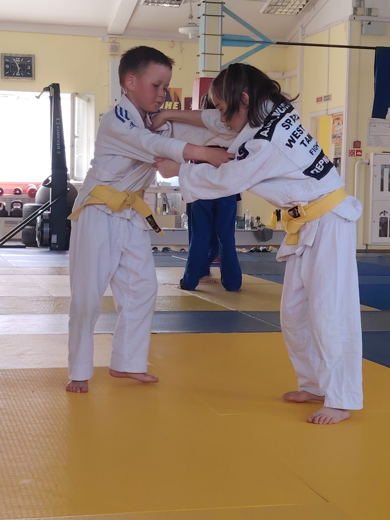 Two children in white judo uniforms with yellow belts are practicing judo on a yellow and blue mat in a gym in Chaikovsky, Russia. One child has their back to the camera with lettering that appears to say "WEST TAM FIGHT" and "ПЕРМЬ". The other child faces the camera. Behind them, there is an exercise machine, some dumbbells, and two people with blue pants. The room is bright, and the window is open.
