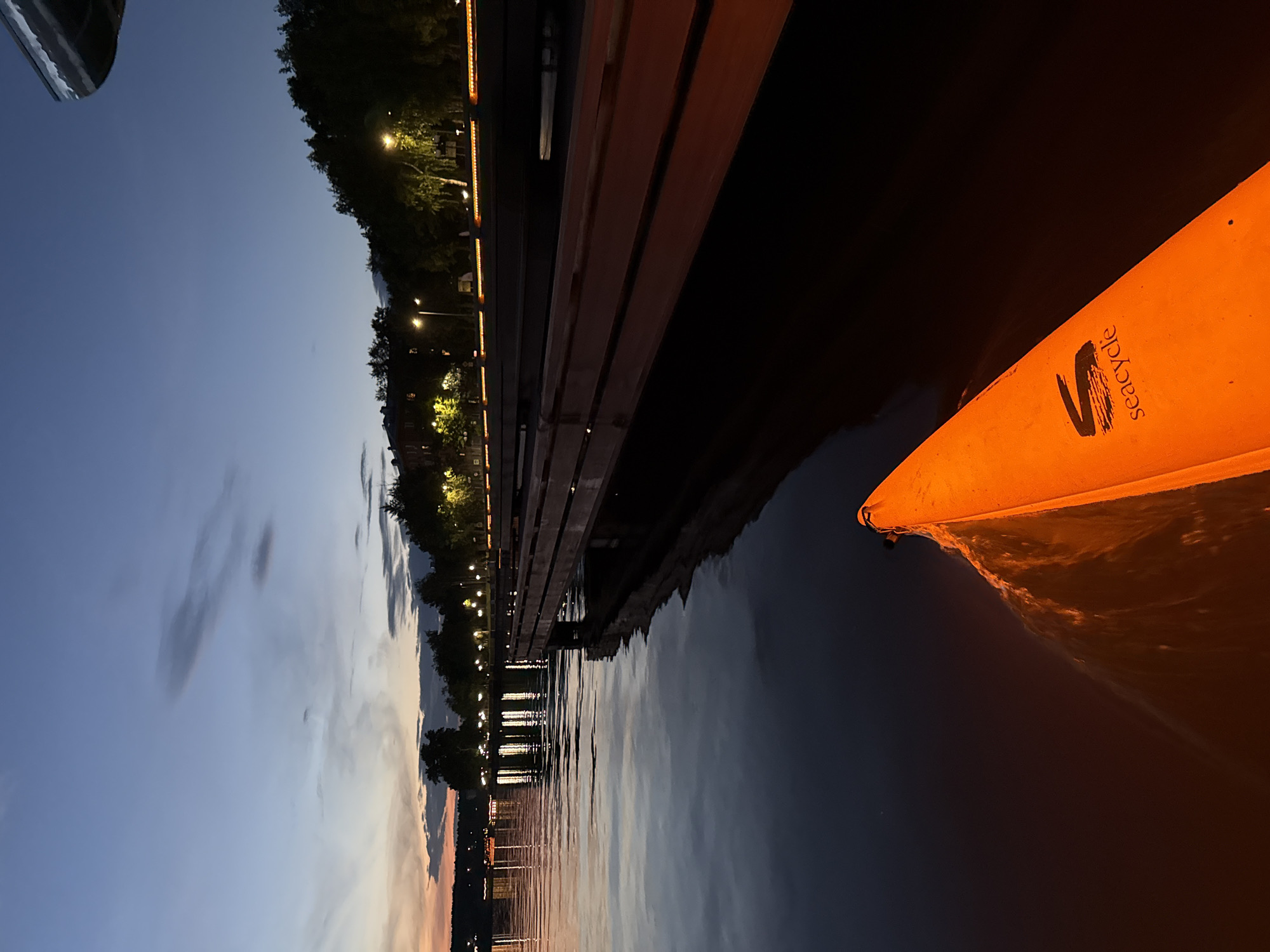 The media file depicts a tranquil waterfront scene at dusk in Votkinsk, Russia. The image is oriented vertically, with the sky occupying the left half and the water, a wooden pier, and an orange object on the right. The sky transitions from a soft blue near the horizon, where hints of sunset colors linger, to a deeper blue at the top. Wispy clouds are visible, adding texture to the twilight sky.
Along the right edge of the sky, a line of trees with illuminated streetlights suggests a populated area bordering the water. The wooden pier runs parallel to the water's edge, its planks a warm reddish-brown. The water is calm, reflecting the lights from the pier and the dusky sky, creating a shimmering, dark surface.
Dominating the right side of the frame is a bright orange object, likely a buoyancy aid or a piece of equipment for a watercraft. The word "seacycle" is visible in black text on this orange object, along with a stylized black graphic. The object appears to be partially submerged or resting on the water, with ripples emanating from it.
The overall mood is peaceful and serene, indicative of the quiet transition from day to night. The lighting suggests it is early evening, with artificial lights beginning to illuminate the scene. The presence of the pier and the implied water activity point towards a recreational area or a dockside.