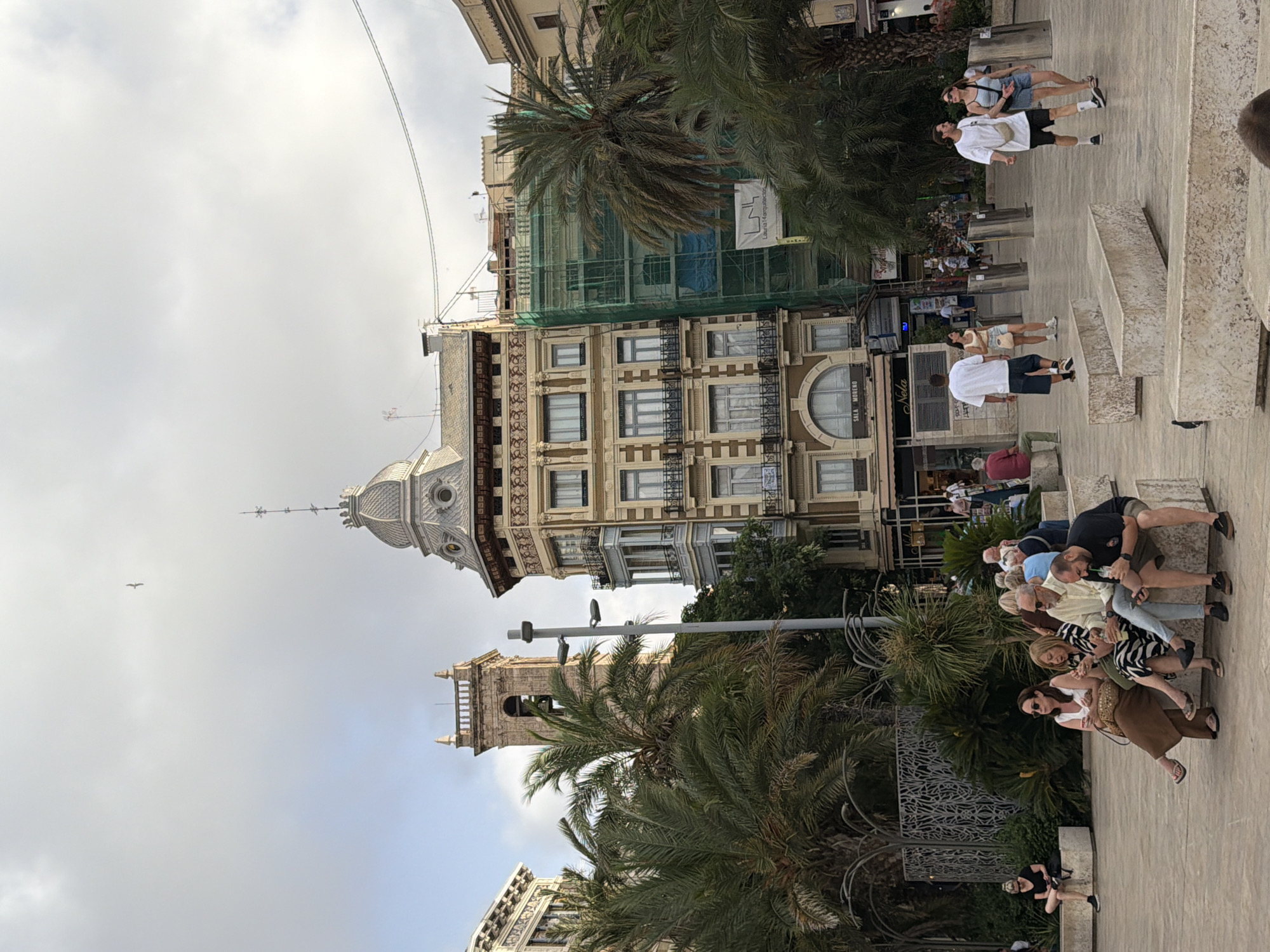 In a city square, people are gathered on benches and walking. In the background, ornate buildings stand tall, one under construction covered in green scaffolding. Palm trees are scattered throughout the scene, and the sky is overcast with a bird in flight. A group of individuals are seated on concrete benches in the foreground. Some are looking at their phones, while others are conversing. A man in a white t-shirt and dark shorts walks with a woman in a blue tank top and denim shorts. Another couple, also in white t-shirts and shorts, walks together. In the distance, more people are visible walking and seated.