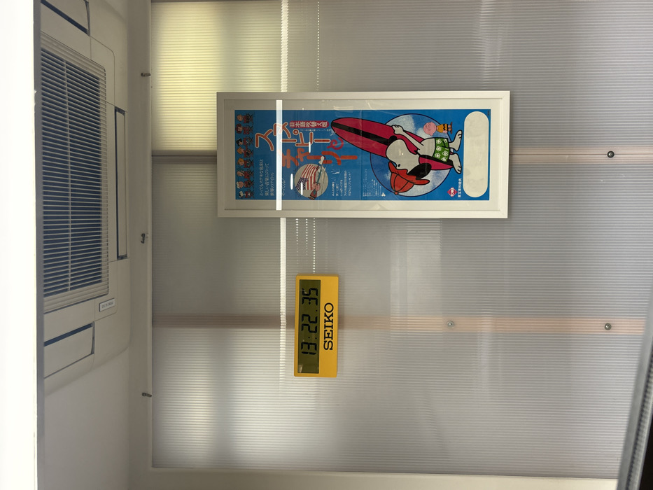 An interior view displays a white ceiling with a rectangular air conditioning vent and a wall composed of frosted, vertically ridged, translucent panels. A yellow rectangular digital clock, branded "SEIKO," is mounted on the left panel, displaying "13:22:35" in black digits. To its right, a white-framed vertical poster is affixed to the wall. The poster depicts the cartoon character Snoopy wearing a red hat and green patterned swim trunks, holding a red surfboard, accompanied by a smaller Peanuts character. The poster's blue background includes cloud designs and prominent Japanese text, such as "スヌーピー" and "東京東和," along with an American flag graphic. Ambient light emanates from behind the translucent panels. No individuals are present. There is no visual information in the image to establish a location in London, United Kingdom; the visible Japanese text on the poster indicates a location in Japan.