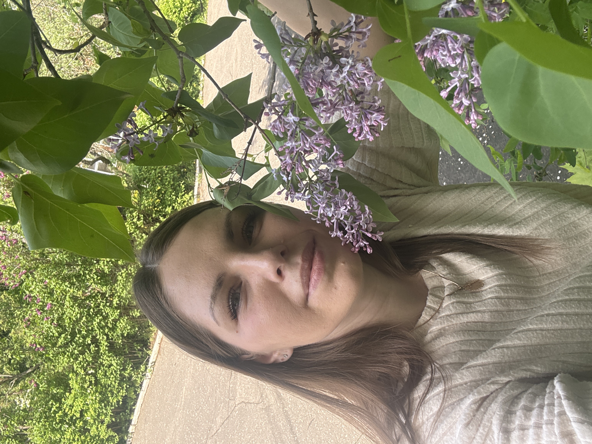 A selfie of a woman, presumably in her early to mid 30s, with medium-length brown hair, fair skin, and light makeup. She is wearing a light-colored textured top and a delicate necklace. She is positioned among flowering lilac branches. The background consists of greenery, what appears to be a building with a light-colored facade, and a paved walkway or road. The image suggests an outdoor environment, likely a park or residential area with blooming lilacs. No evidence in the image indicates a specific location in Shevlyakovo, Russia.
