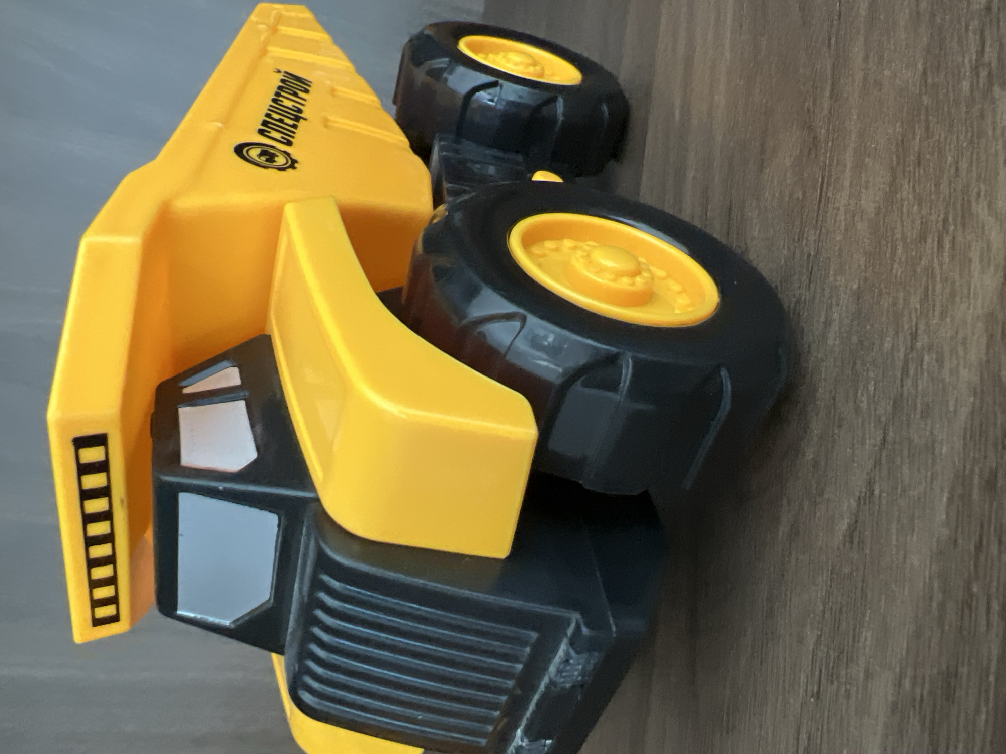 A yellow toy dump truck is positioned on a wooden surface, likely a table or floor, against a gray background. The truck is primarily yellow and black, with black wheels that have yellow rims. A logo with the word "СПЕЦСТРОЙ" in Russian is visible on the side of the truck's bed. The truck is the sole subject of the image. The scene is static and shows no activity. The location is unknown, and there is no indication of it being in Минзаг, Russia.