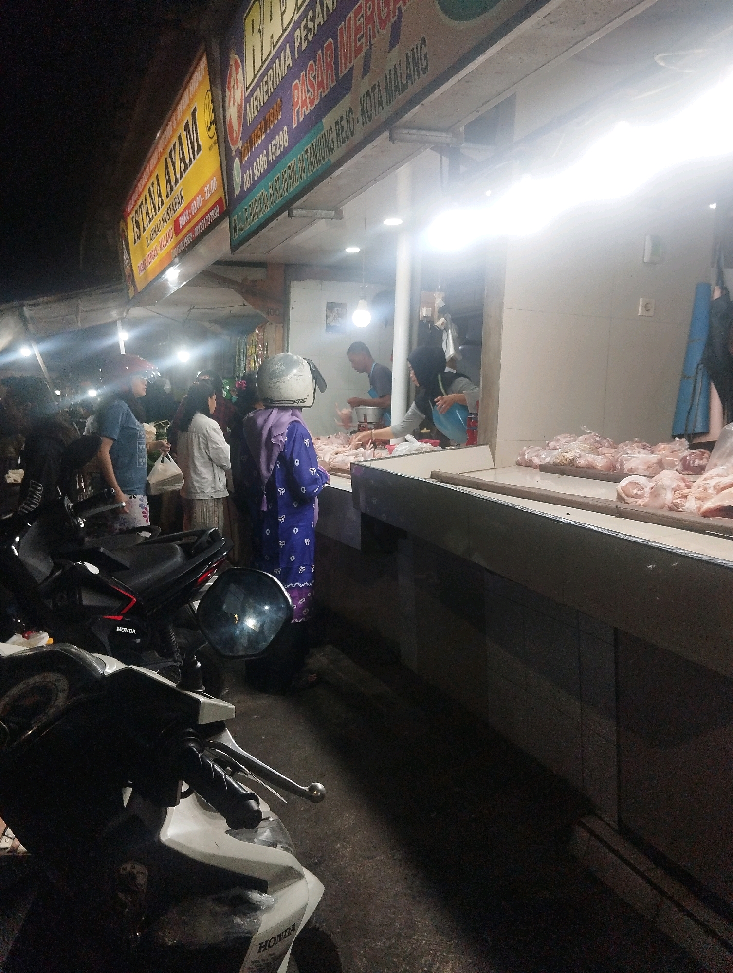 The image captures a night scene at a busy market in Malang, Indonesia, specifically at a chicken stall. In the foreground, a white scooter is parked, partially obscuring the view. The stall itself is brightly lit by fluorescent lights, illuminating a large display of raw chicken cuts arranged on a counter. Several people are visible around the stall. A woman wearing a purple patterned outfit and a white helmet stands facing away from the camera, while another person behind the counter appears to be serving a customer. To the left of the stall, a man in a blue shirt and a woman in a white jacket are also present, with the woman holding a white plastic bag. In the background, another stall with yellow and purple signage advertising "ISTANA AYAM" and "PASAR MERGO" can be seen, further indicating the location. The overall atmosphere suggests a bustling night market environment.