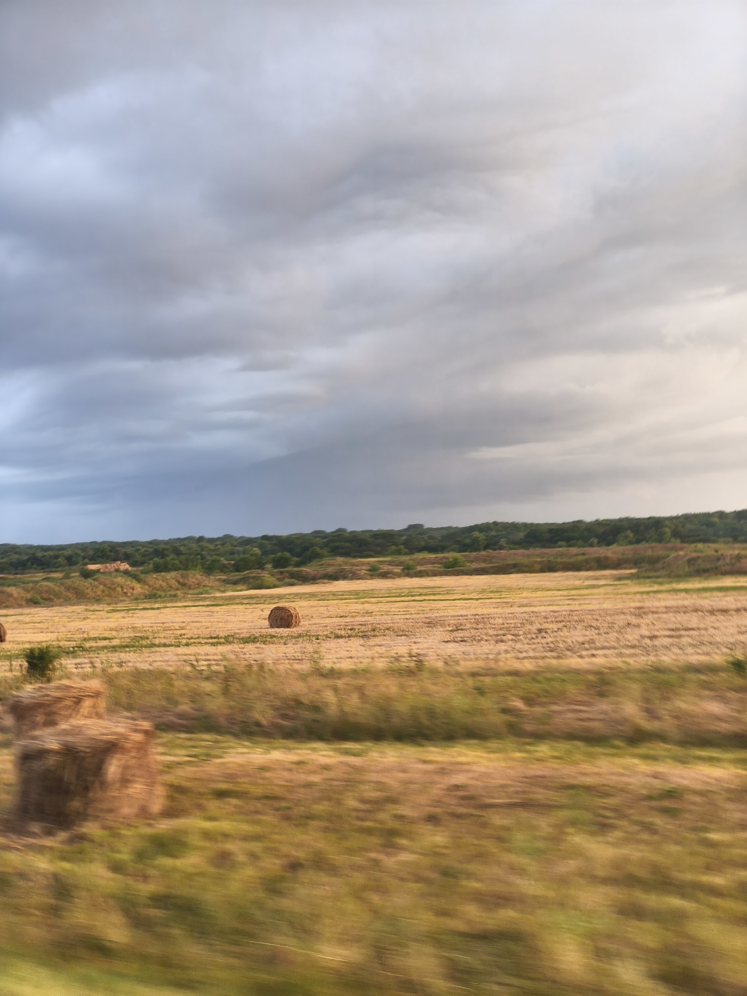 The image depicts a rural landscape with a field of harvested crops and several hay bales under a cloudy sky. The scene is characterized by a motion blur effect, suggesting the photograph was taken from a moving vehicle. In the mid-ground, a single, round hay bale is visible in a harvested field. To the left, a stack of rectangular hay bales is present. The background features a dense line of green trees bordering the field. The sky is filled with a mix of light and dark gray clouds, with some patches of lighter sky towards the right. There are no people or animals visible in the image, and no specific identifiable landmarks of Тоннельный, Russia.
