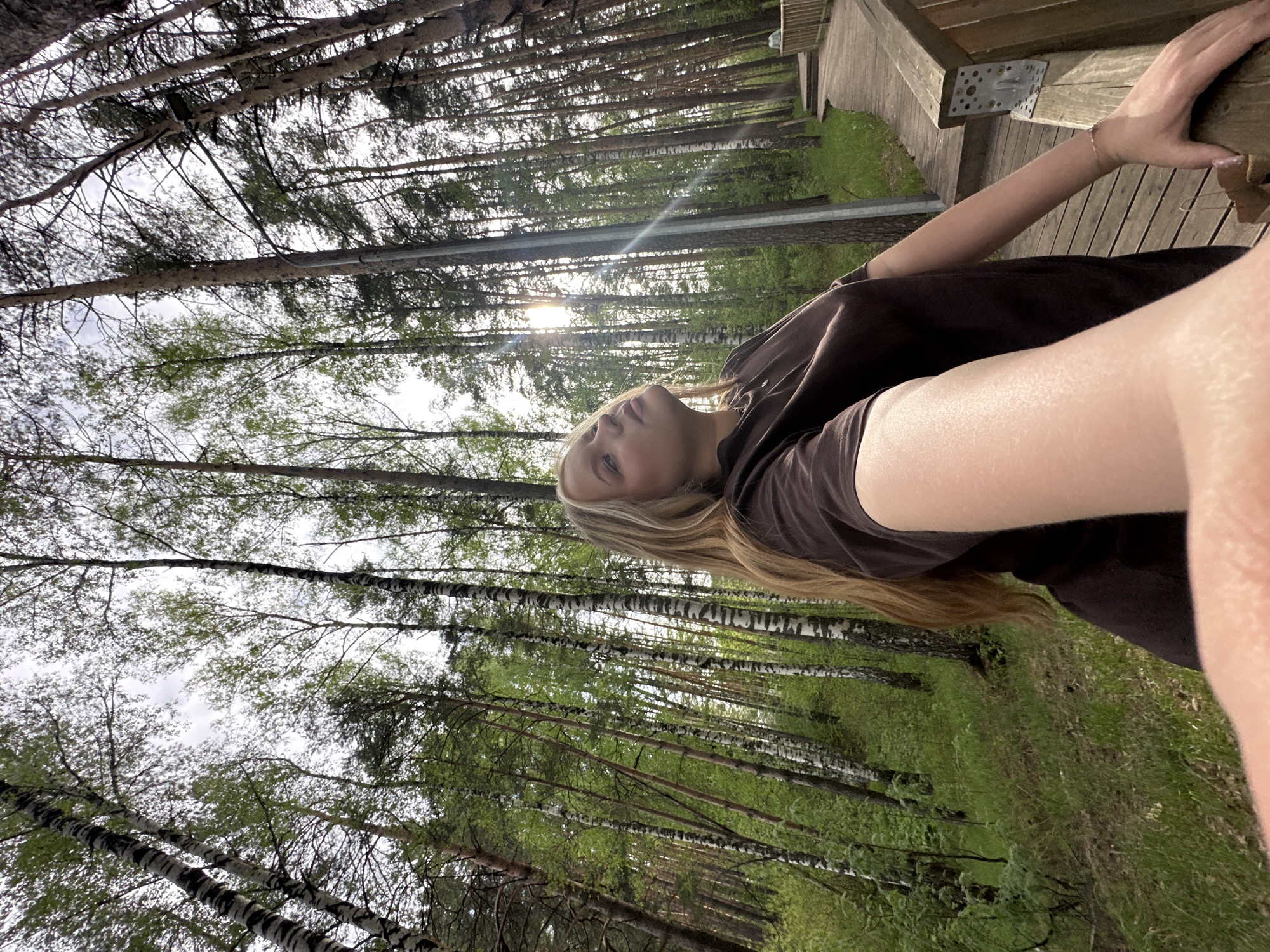 A person is outdoors in a forest landscape in Chaikovsky, Russia. The person is close to the edge of a wooden deck, visible in the upper right of the frame. The forest consists primarily of tall, slender trees that appear to be a mix of pine and birch, characterized by their white bark. Sunlight filters through the tree branches. The undergrowth is lush green vegetation. The person is fair-skinned with long, blonde hair and is wearing a brown top. One of their arms is extended, with their hand resting on the wooden railing of the deck.