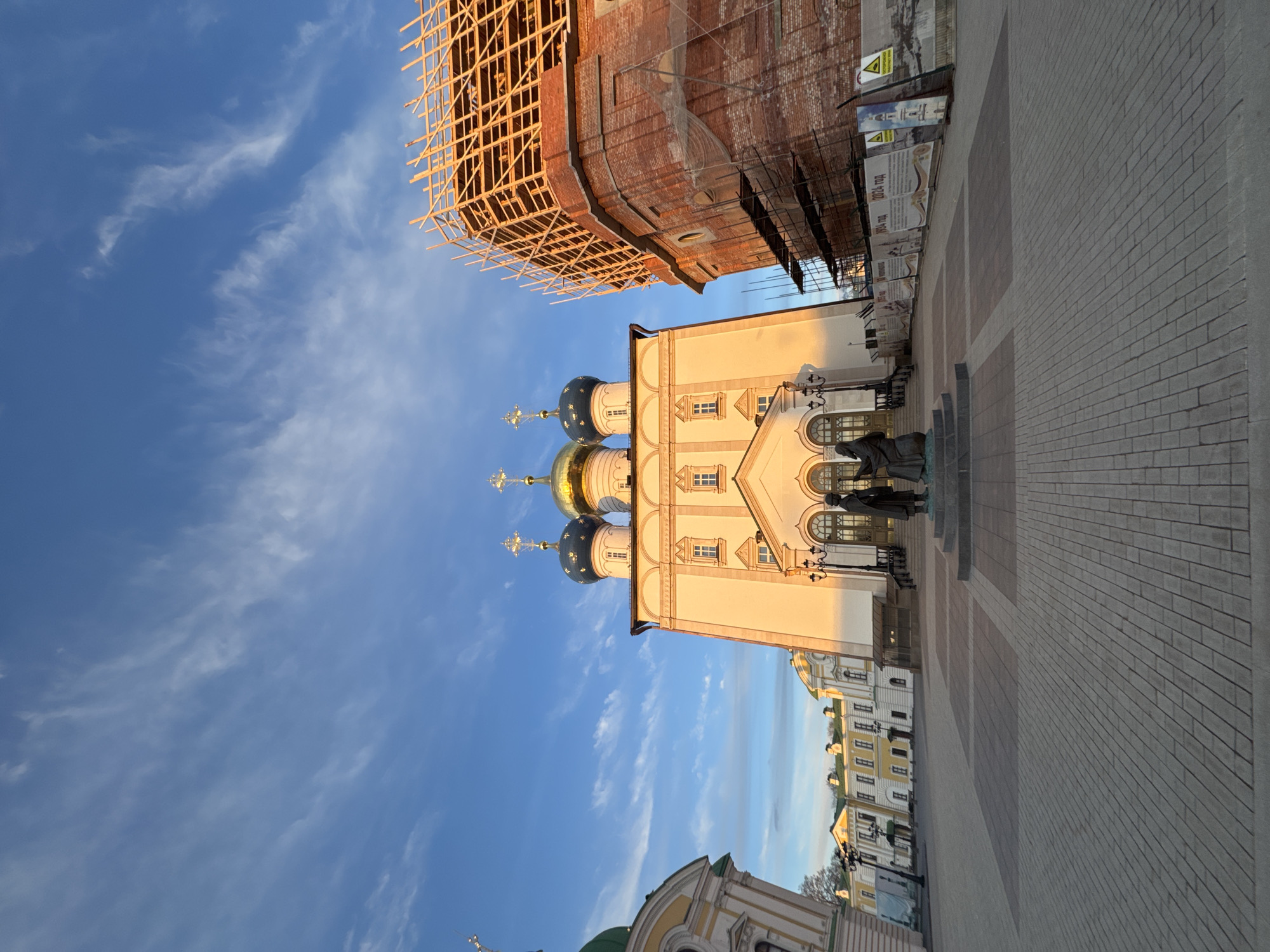 The image shows a view in Tver, Russia, featuring the partly-constructed Transfiguration Cathedral, along with the restored White Trinity Church. The cathedral is a large brick structure under construction, with scaffolding visible on top. Adjacent to the cathedral is the cream-colored White Trinity Church with four blue domes topped with golden crosses. A statue stands in front of the church on a stone pedestal. The ground is paved with grey and tan bricks. In the background, other buildings with yellow facades and dark roofs can be seen. The sky is blue with scattered white clouds.