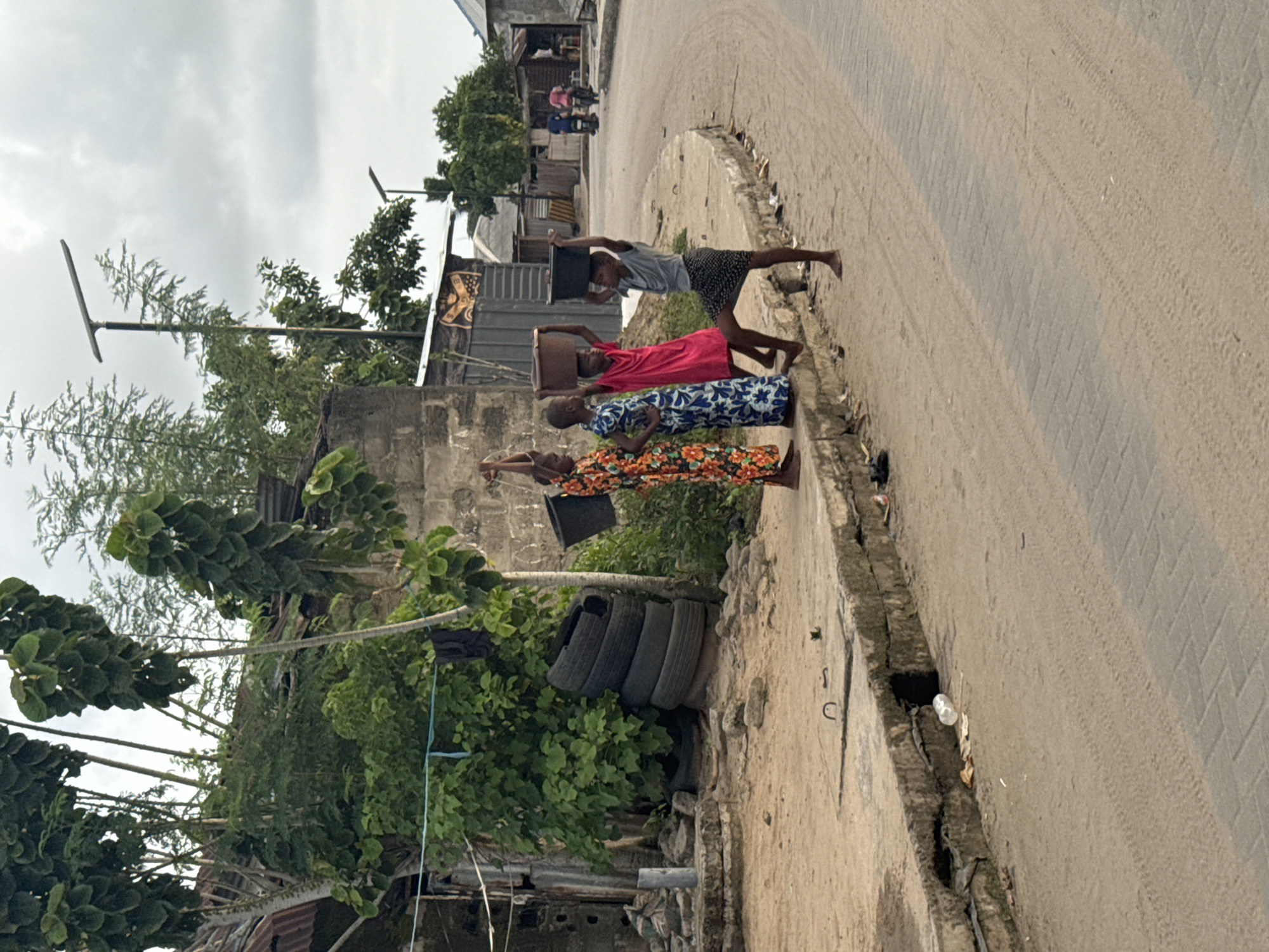 Three people are walking barefoot along a road in Badagry, Nigeria, each carrying a container on their head. The first person is wearing an orange and floral pattern dress, the second is wearing a blue and floral pattern dress, and the third person is wearing a pink dress. A fourth person is walking further down the road, wearing a grey shirt and black and white shorts, also carrying a container on their head. The road is made of asphalt and is lined with trees and buildings. There are several old car tires piled up on the side of the road. Overhead cables and an overcast sky are visible.