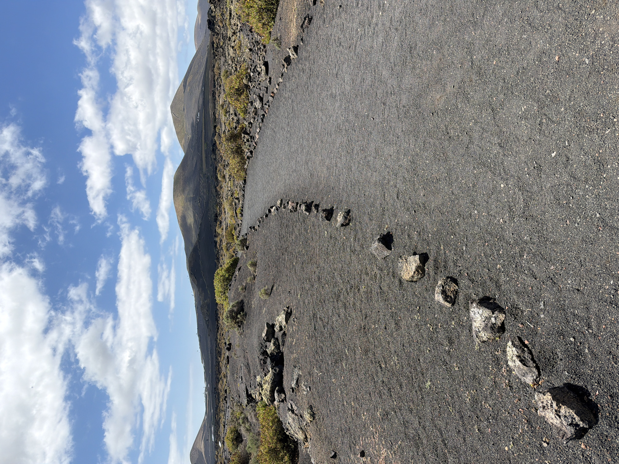 The image captures a serene, rugged volcanic landscape under a clear sky. A narrow, winding path of dark grey gravel or volcanic ash dominates the foreground and midground, leading into the distance. This path is delineated on both sides by irregularly placed, dark rocks, suggesting a marked trail for walkers.
To the left and right of the path, the ground is covered in similar dark, granular material, interspersed with patches of sparse, hardy green and yellowish vegetation, typical of arid, volcanic environments. In the midground and background, rolling hills and mountains rise, their slopes also appearing dark and rocky, indicative of volcanic origin.
The sky above is bright blue with scattered white, fluffy clouds, suggesting a clear and sunny day. The lighting indicates it is daytime, possibly late morning or early afternoon, with no harsh shadows. There are no people, animals, or visible text in the scene. The overall impression is one of vastness and natural beauty in a unique geological setting, consistent with the volcanic landscapes found around Tías, Lanzarote, Spain.