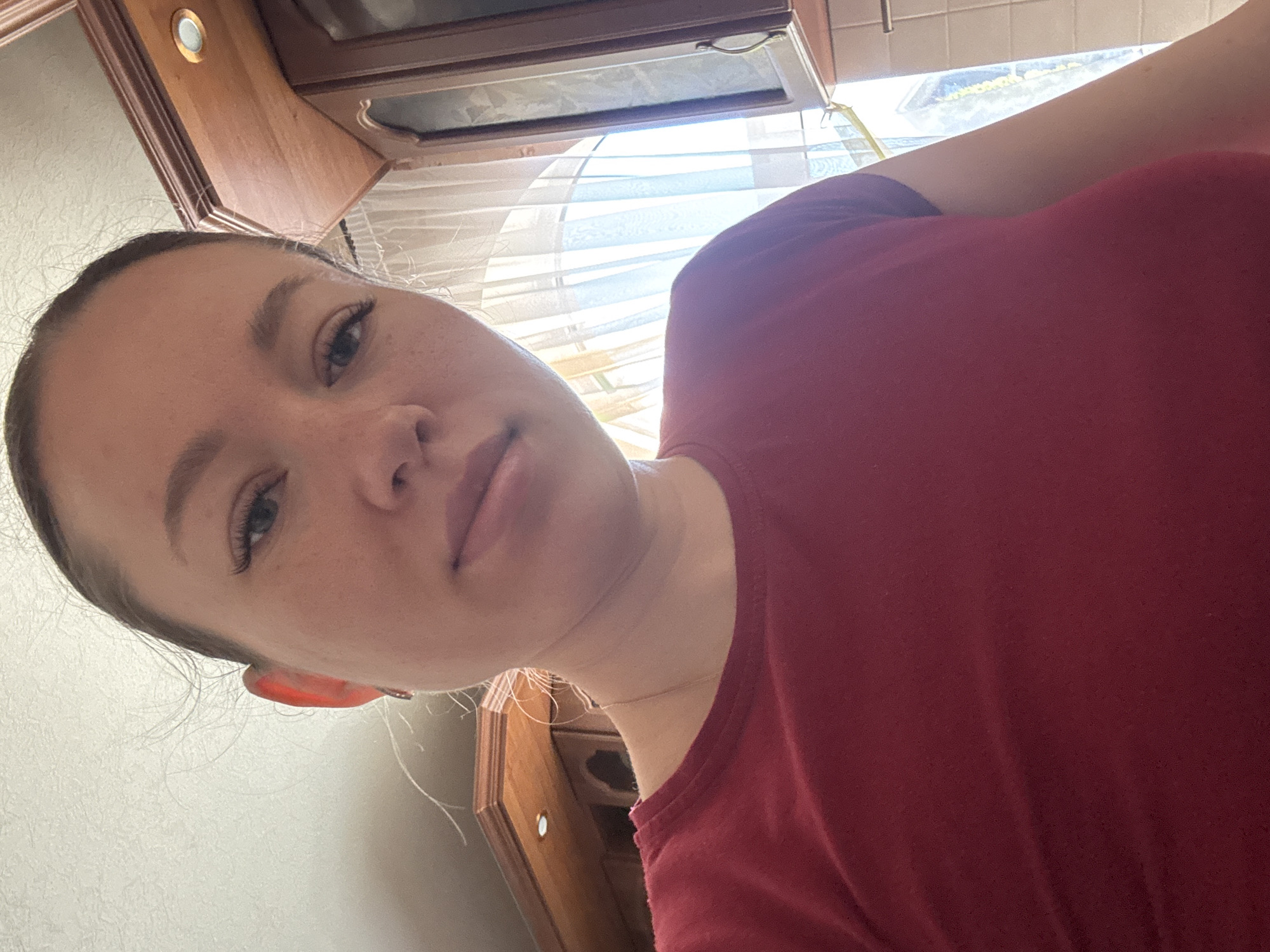 A selfie photograph taken in what appears to be a kitchen. A person with fair skin and dark hair, likely female, is centered. She wears a maroon t-shirt. The background includes kitchen cabinets and a window with light filtering through white curtains. The location in the city of Ферма, Russia is not discernible from this image.