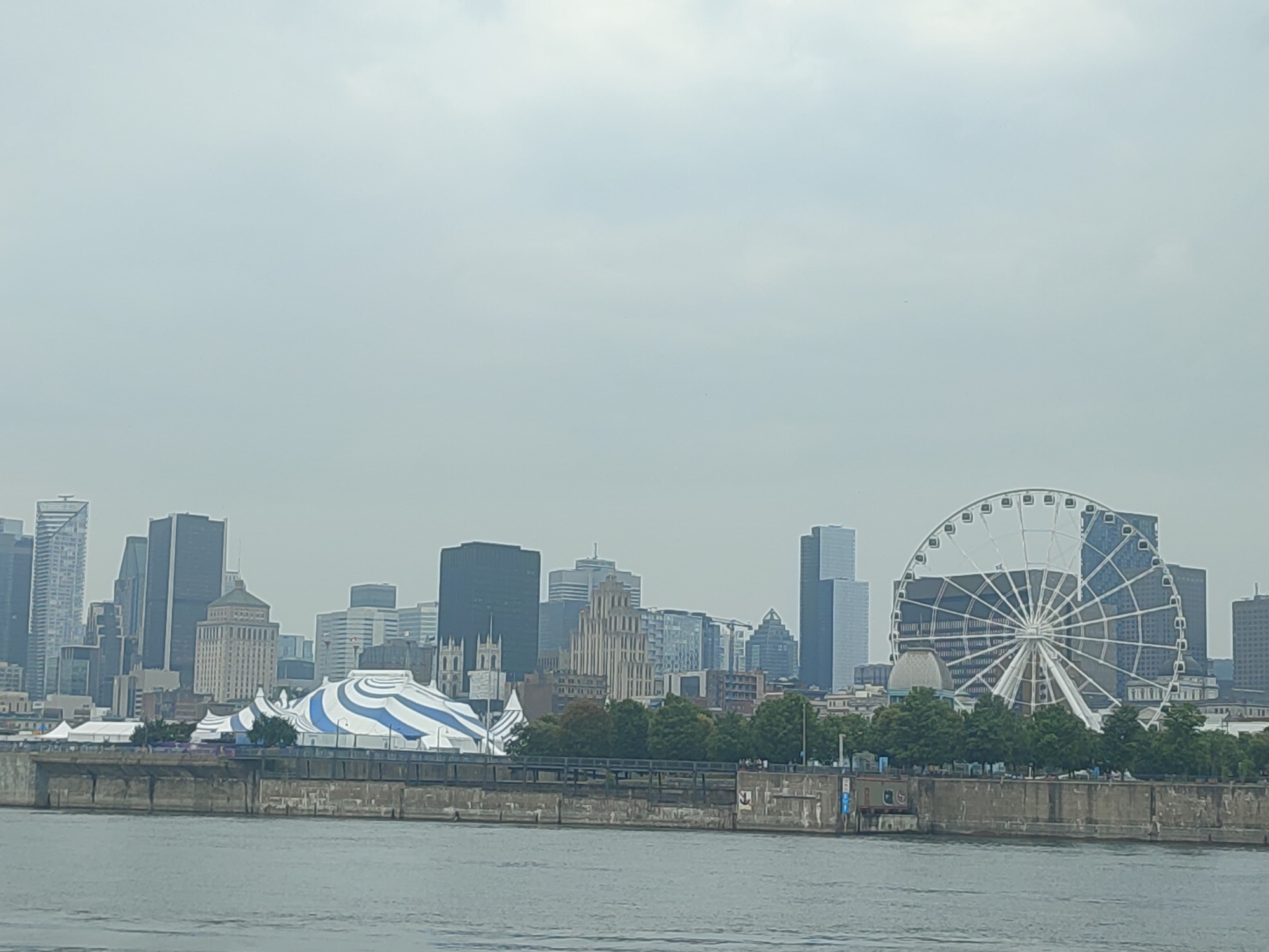 This is a daytime, wide shot of the Montreal skyline from across the water on an overcast day. The foreground shows a body of water. Behind the water is a concrete embankment with some metal structures and a wooden walkway. Beyond the embankment, the Montreal skyline rises. Prominent features include a large white and blue striped tent or circus structure, a large Ferris wheel, and numerous skyscrapers of varying heights and architectural styles. Trees are visible along the waterfront in front of the skyline. The sky is heavily clouded and gray. There are no visible people or animals in the image.