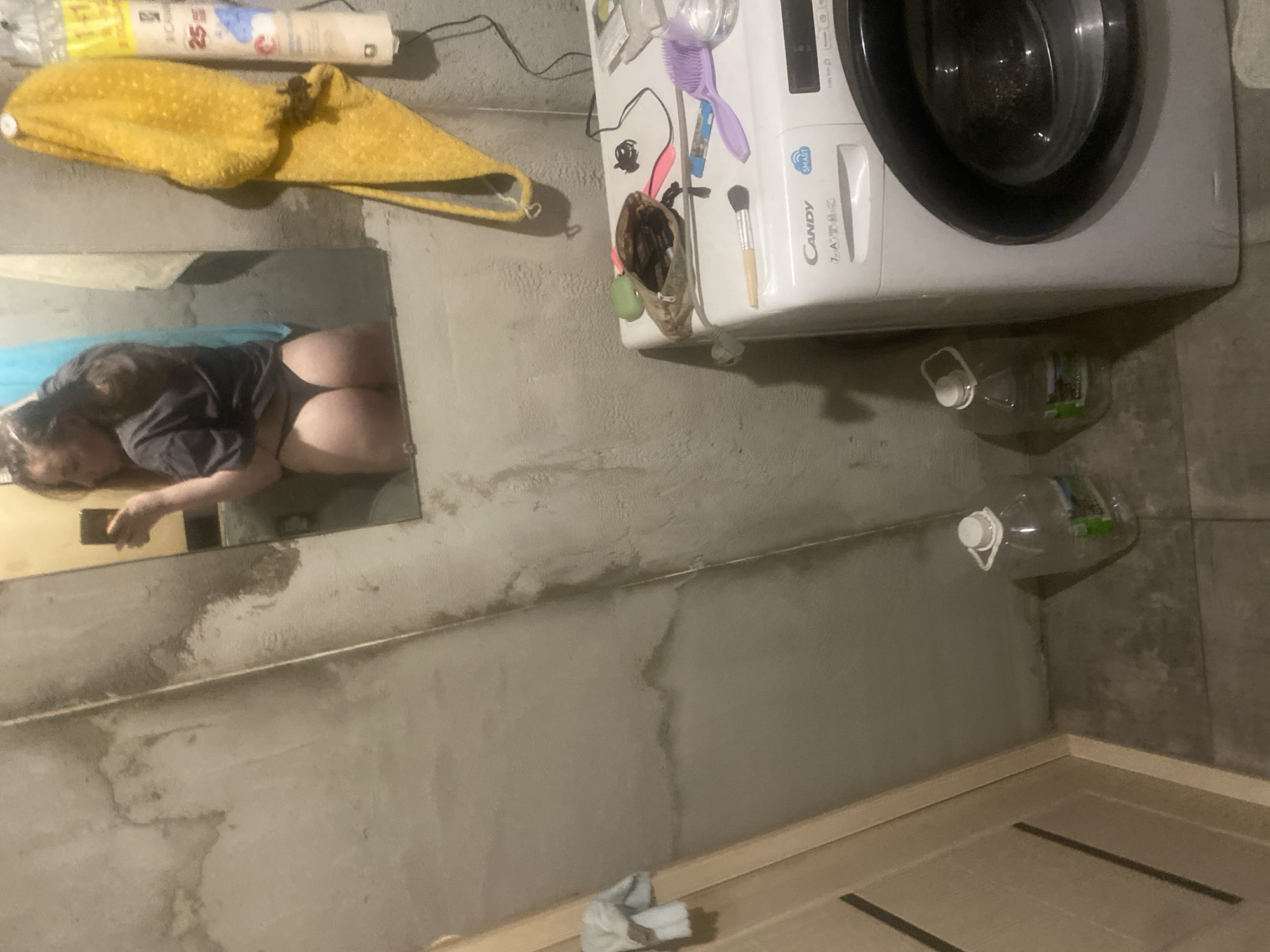 In an indoor environment with a concrete-like wall, a woman is seen in a mirror's reflection, holding a smartphone. She is wearing a dark t-shirt and underwear. To the right of the mirror, a yellow towel hangs. In the foreground, a white washing machine is visible, with various items on top, including a makeup bag, a brush, and a small blue object. Two large clear plastic bottles with white caps are on the floor to the left of the washing machine. A wooden doorframe is present on the left side of the image. The overall scene appears to be a utilitarian space, possibly a bathroom or laundry area.