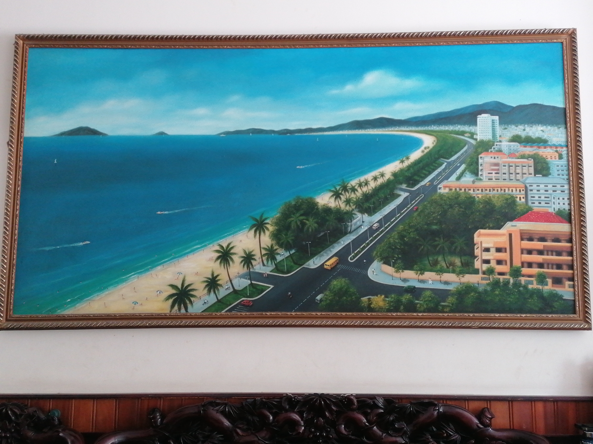 The image is a framed painting depicting a coastal scene. The painting features a wide blue bay with several small islands in the distance. A sandy beach runs along the coast, lined with palm trees and a road with vehicular traffic. Behind the road are buildings, including a prominent high-rise, and mountains in the background. The scene likely depicts the Nha Trang Ward in Vietnam. Below the framed painting is a decorative, dark-colored wooden surface.