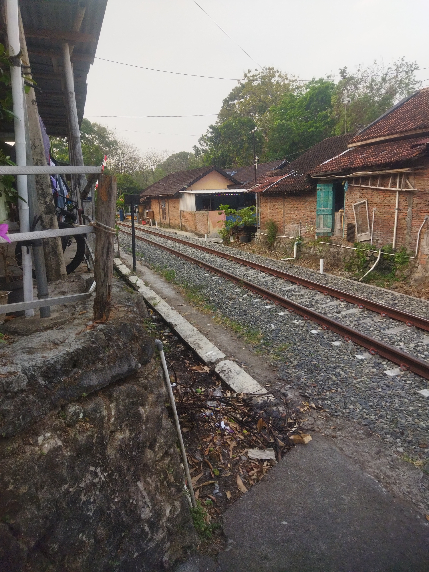 This image depicts a railway track running alongside a row of brick houses in a rural area, likely in Wonogiri, Indonesia. The sky is overcast and appears to be early morning or late afternoon. The foreground features a weathered stone wall and a concrete path next to the tracks, which are covered in gravel and ballast. Debris, including dried leaves and twigs, has accumulated along the edge of the track. To the left, a covered porch with metal supports and corrugated roofing is visible, with laundry hanging on a line. A bicycle is propped against a wooden post. The houses have red tile roofs and brick walls, with some featuring green wooden shutters and windows. Trees and foliage can be seen in the background. The scene suggests a quiet, residential area adjacent to active railway infrastructure. There are no people visible in the frame.