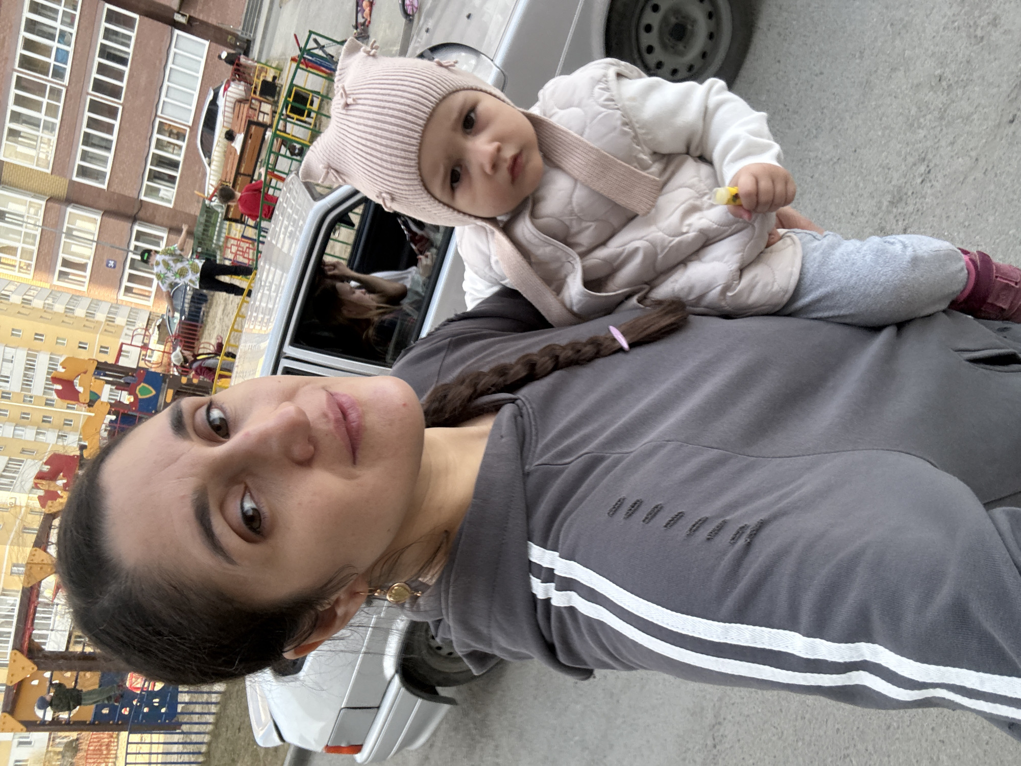 The image is a vertically oriented selfie, captured from a tilted perspective, showing a woman and a baby outdoors in what appears to be a residential area of Tobolsk, Russia.
In the foreground, the woman has dark hair styled in a braid, adorned with a pink hair tie. She wears a grey hooded sweatshirt featuring white stripes on the arm and subtle distressed details on the chest. A golden-colored earring is visible on her right ear as she looks towards the camera with a neutral to slightly smiling expression.
Resting on the woman's shoulder or lap is a baby, dressed in a light pink knitted hat with ear-like protrusions, a white long-sleeved top, a quilted light-colored vest, light grey pants, and magenta-pink shoes. The baby holds a small yellow object in its right hand and gazes directly at the camera with a curious or serious expression.
The background reveals a typical residential courtyard with multi-story apartment buildings; one is reddish-brown, and another is yellow, both featuring numerous windows. A vibrant children's playground with red, yellow, green, and blue equipment is prominent. A silver car with an open passenger door is parked beside the subjects, reflecting a person. Several other individuals are visible in the background, including a person in a green cap and a pineapple-patterned shirt, and another in a red top near the playground. The ground is paved with asphalt or concrete. The scene takes place during the daytime, under bright, possibly overcast conditions. A blue street sign displaying the number "34" is visible on one of the buildings.