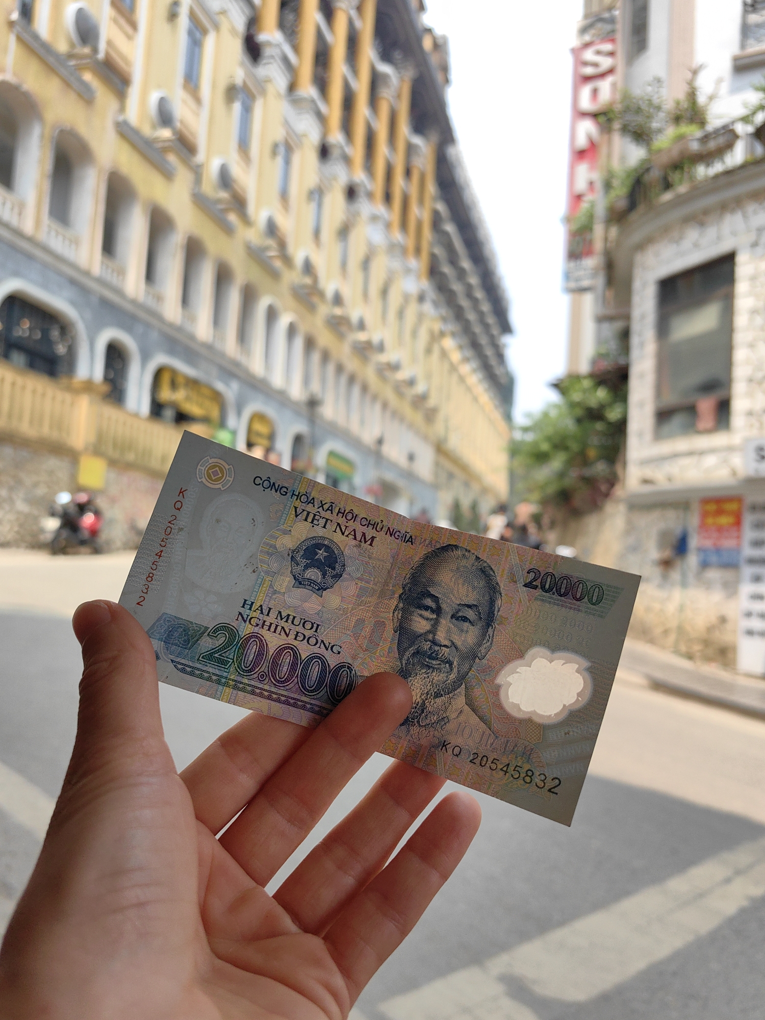 A light-skinned human hand is centrally positioned, holding a 20,000 Vietnamese Dong banknote in the foreground. The banknote's obverse is visible, featuring a portrait of Ho Chi Minh on the right, the text "CỘNG HÒA XÃ HỘI CHỦ NGHĨA VIỆT NAM" at the top, and "HAI MƯƠI NGHÌN ĐỒNG" below the national emblem. The serial number "KQ 20545832" is printed vertically on the left side. The note exhibits blue, green, and pink coloration.
The background is an out-of-focus street scene in the city of Sa Pa, Vietnam, illuminated by natural daylight. To the left, a multi-story building with a yellow facade, numerous arched windows, decorative columns, and visible air conditioning units lines the thoroughfare. On the right, a corner building displays a vertical red sign with white lettering, partially obscured but resembling "SAPA" or "SUN," and features small planters with greenery on its balconies. A paved road surface occupies the lower and mid-ground. A blurred red motorcycle is discernible on the left side of the road in the mid-distance. Shadows on the road indicate an overhead light source. No distinct individuals are clearly identifiable beyond the hand holding the currency.