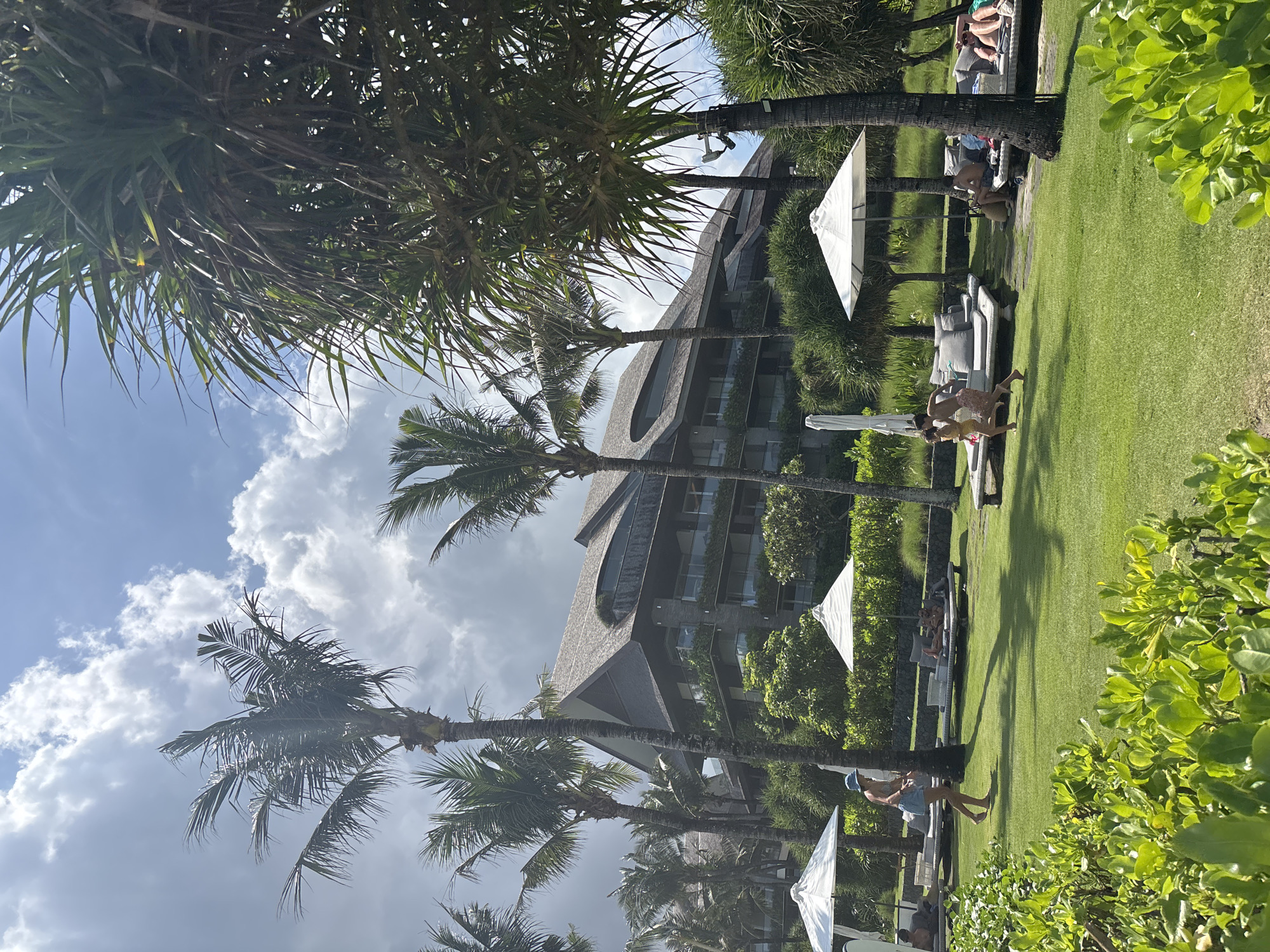 The image depicts a resort area in North Kuta, Indonesia. A multi-story building with a distinctive roof design is visible in the background, partially obscured by palm trees and other tropical vegetation. In the foreground, there is a well-manicured lawn with lounge chairs and white umbrellas. Several people are relaxing on the chairs, and others are walking around. The sky is partly cloudy with blue patches visible. The lighting suggests it is daytime. Overall, the scene conveys a relaxing, tropical vacation atmosphere.