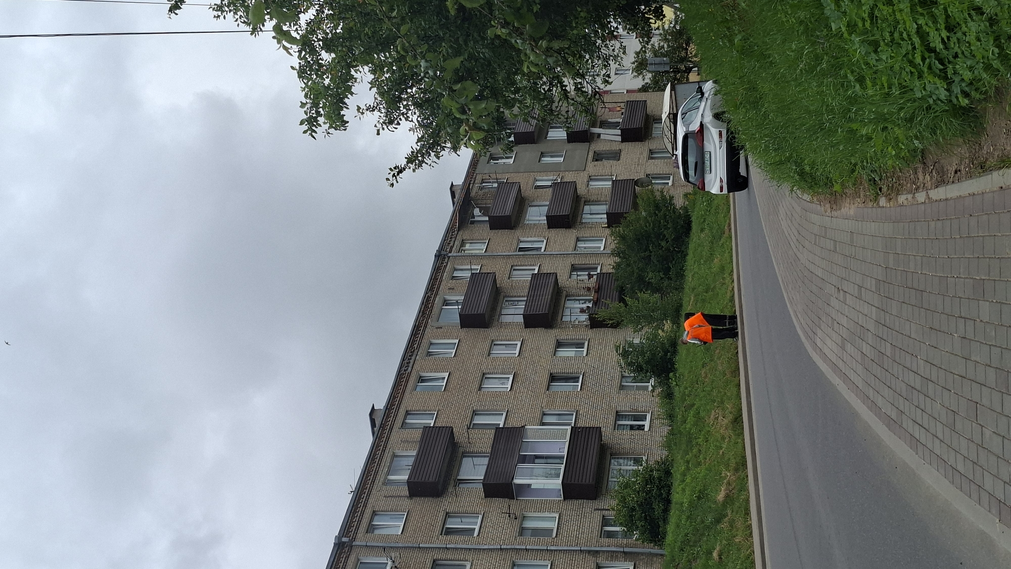 The image captures a street scene in Smalyavichy, Belarus, under an overcast sky. A multi-story apartment building, constructed with light-colored brick, dominates the background. The building features numerous windows and dark brown balconies. In the foreground, a paved sidewalk runs alongside a road. Two individuals, wearing bright orange safety vests over dark clothing, are standing near the edge of the road and the grassy embankment. Their backs are to the camera, and they appear to be engaged in an activity on the ground. A white hatchback car is parked on the right side of the road, facing away from the viewer. Lush green grass covers the embankment, and trees with dense foliage are visible on the right side of the frame, partially obscuring the building. A single bird is visible in the upper part of the sky.