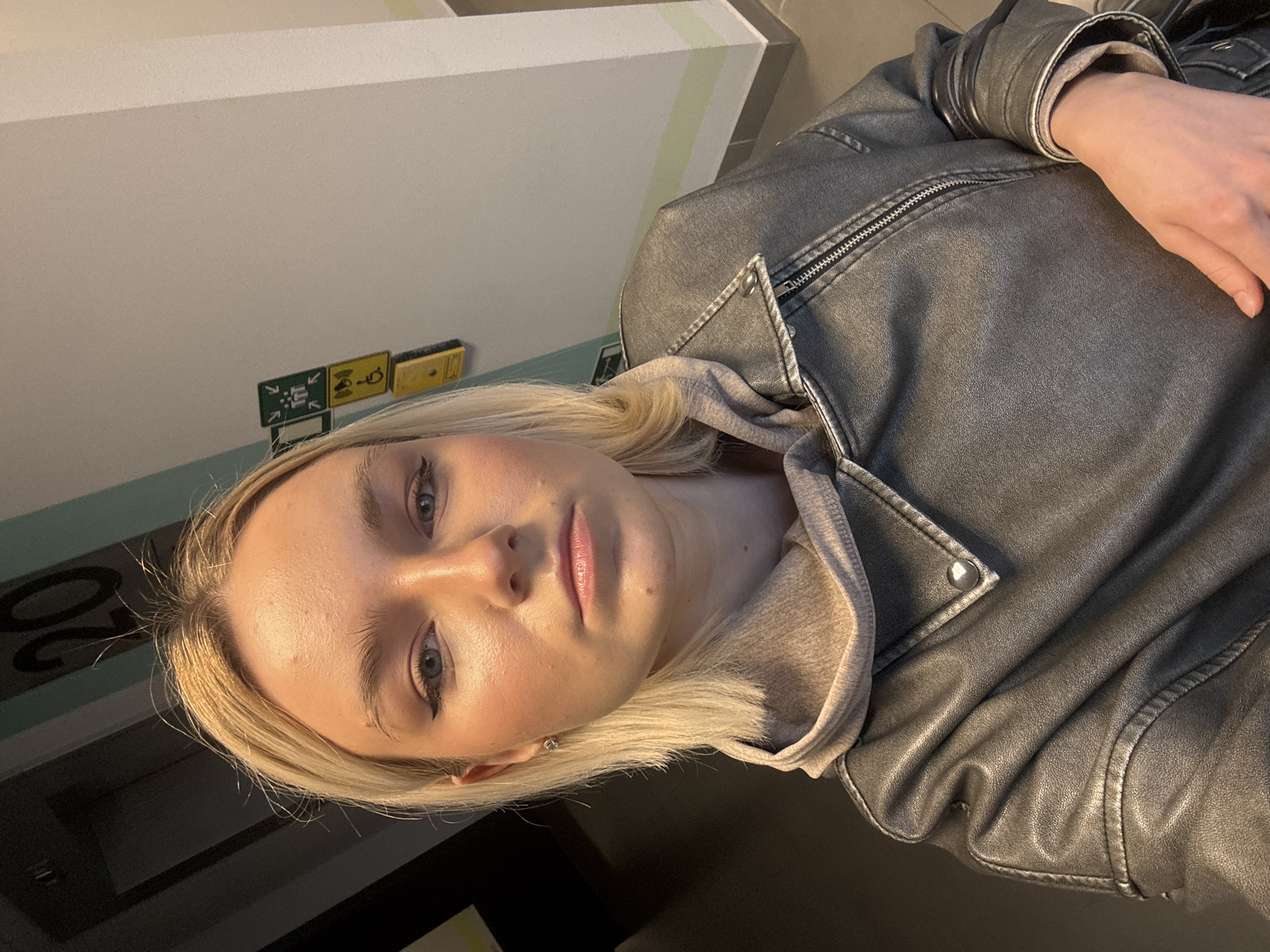 A fair-skinned individual with light blonde hair is shown in a close-up, slightly angled shot. The person is wearing a leather jacket over a hooded top. The background features a wall with a combination of white, light green, and dark gray segments, and some small, unclear signs in yellow and green.