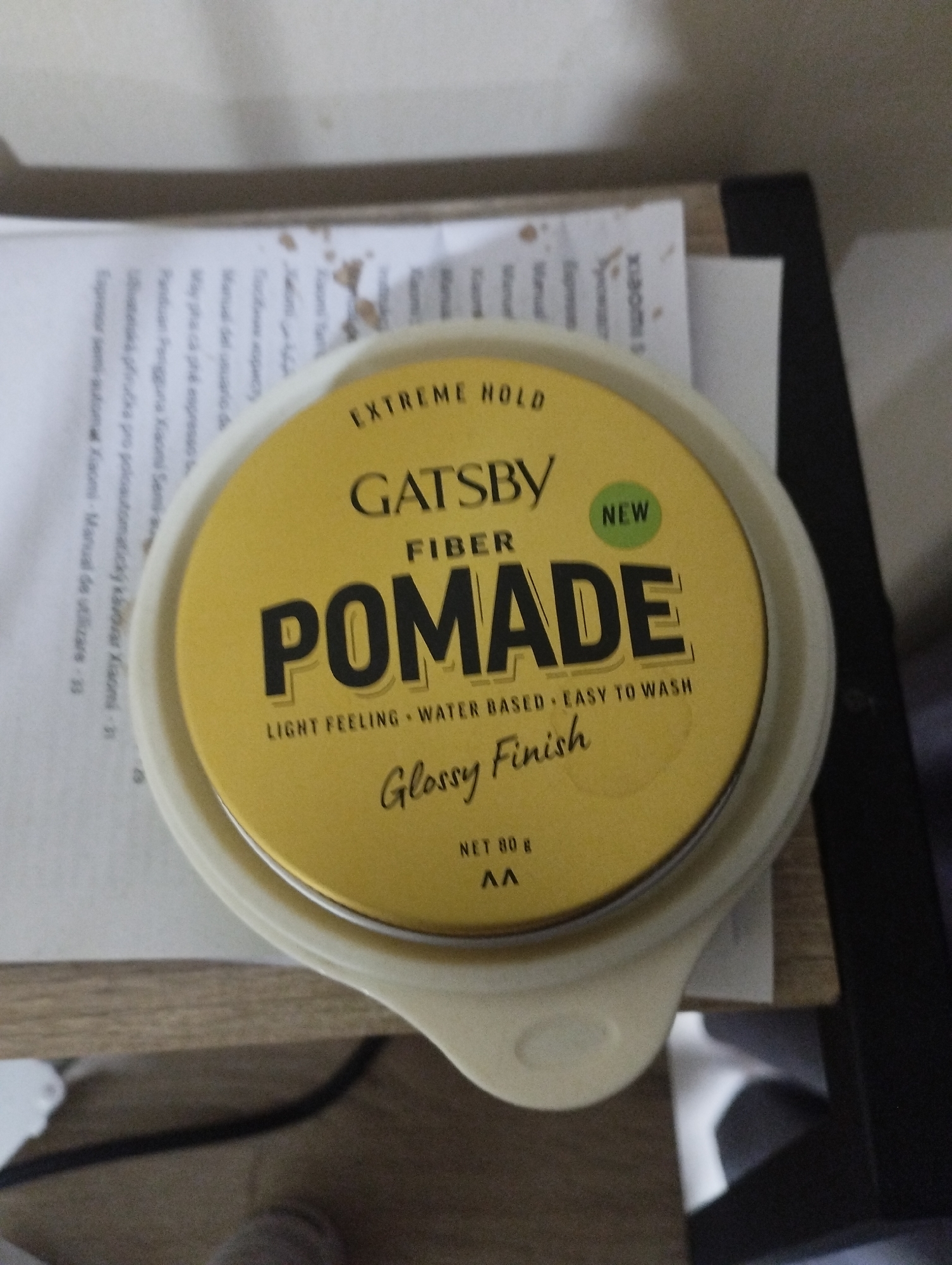 The image shows a container of Gatsby Fiber Pomade with "Extreme Hold" and "Glossy Finish" on top of what appears to be a manual, possibly for a Xiaomi product. The surrounding environment suggests an indoor setting, possibly a desk or table. The location is not discernible from the image. There are no people visible in the picture. The overall presentation gives a casual, almost snapshot-like quality. The presence of the manual indicates some degree of organization but not necessarily tidiness.