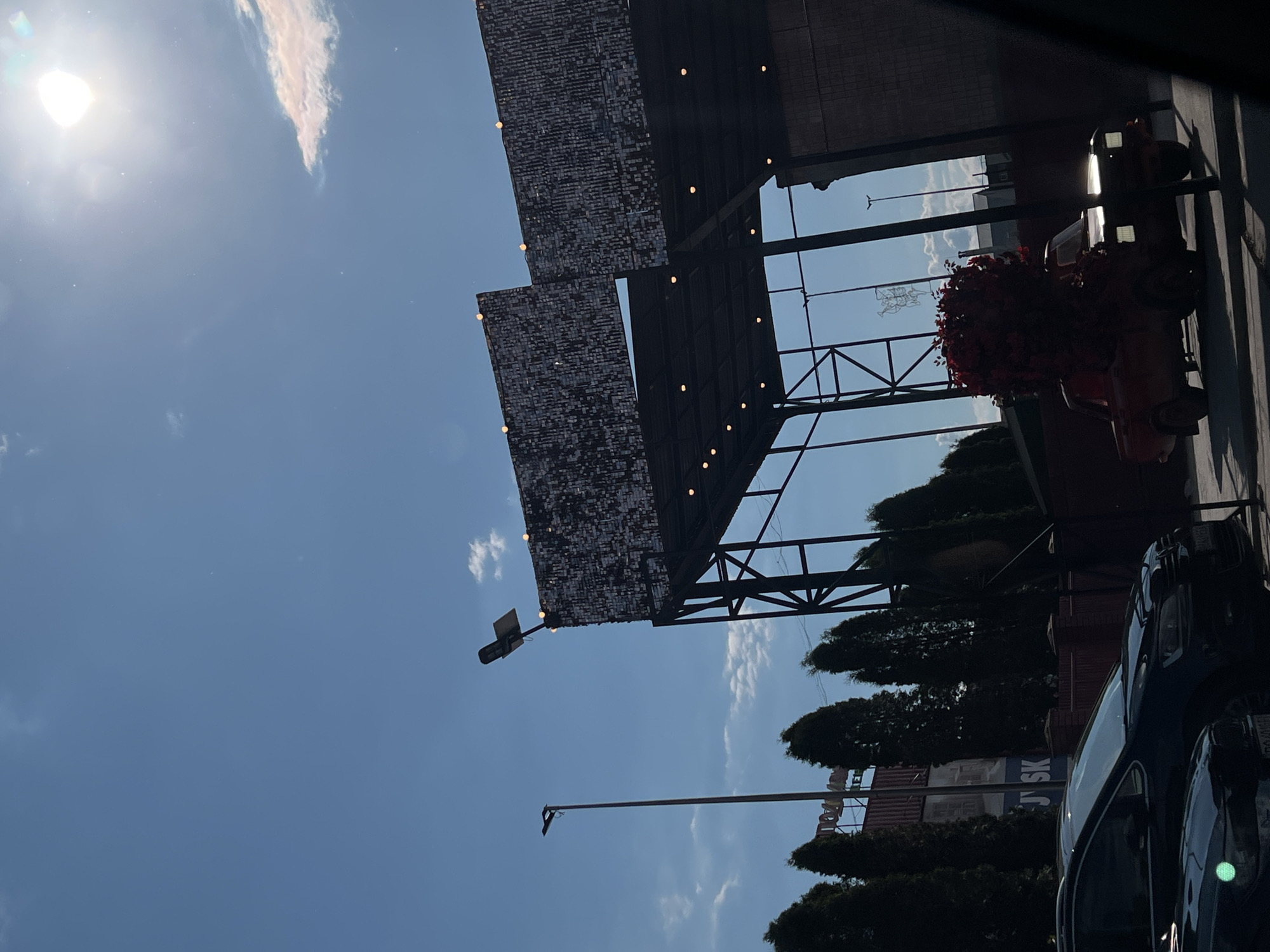 The image depicts a sunlit outdoor scene in Ternopil, Ukraine, featuring a prominent industrial-style structure with large, sequined billboards. A tall street lamp stands to the left of the structure, and a line of evergreen trees runs behind it. In the foreground, the top of two dark-colored cars is visible. To the right, a red vintage car is adorned with a large, red floral arrangement on its hood and roof. The sky is blue with scattered clouds and bright sunlight, which causes lens flare in the upper right corner. The lower portion of the image is shadowed.
