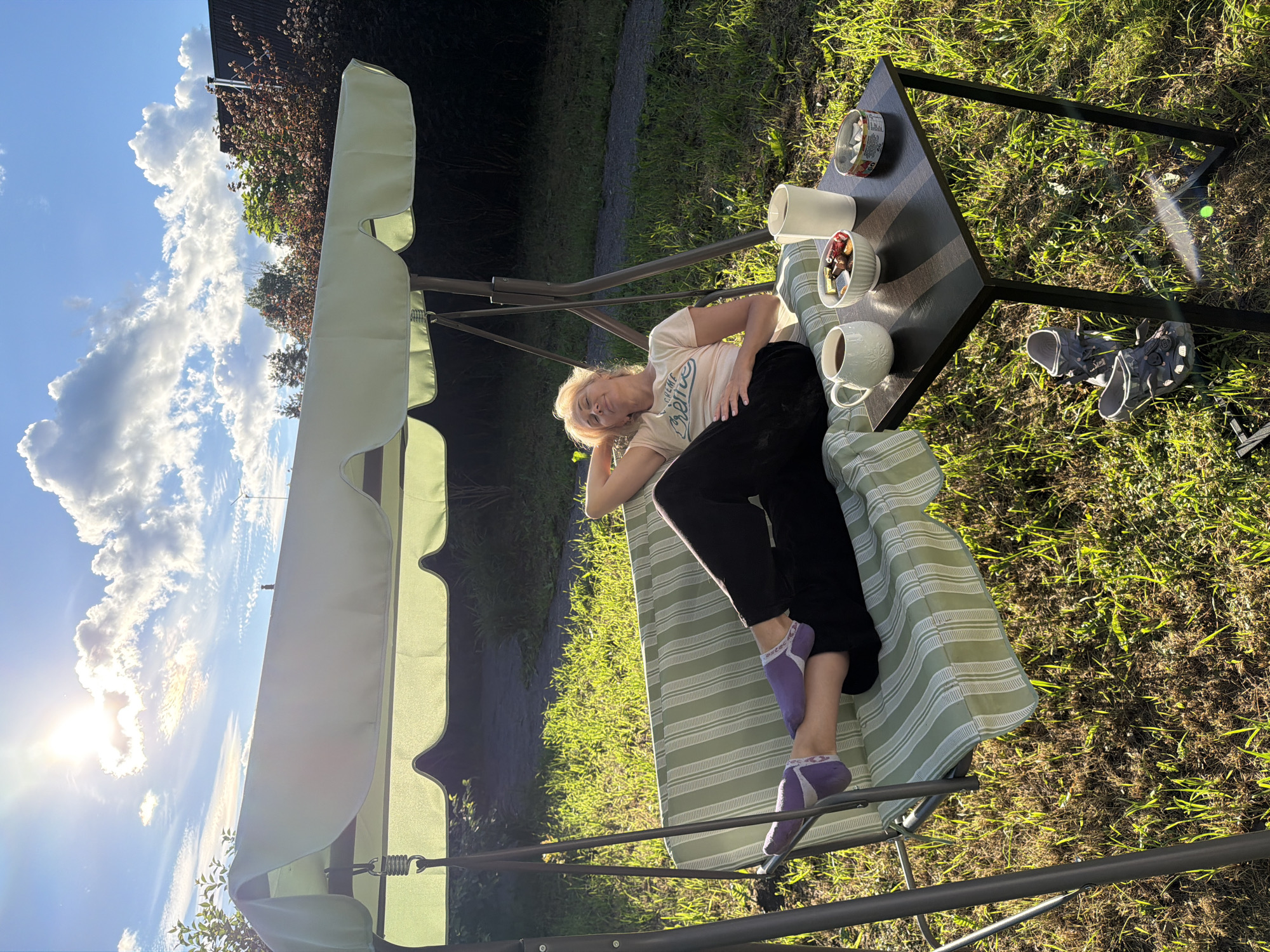 A woman is relaxing on a green and white striped swing seat in a backyard in Steklyannyi, Russia. She is wearing a light-colored t-shirt, black pants, and purple socks. Her head is propped up on her hands, and her eyes are closed. A small black table is next to the swing, with two mugs of coffee, a bowl of candy, and a small tin can on it. A pair of grey Crocs are on the ground next to the table. The sun is shining brightly, casting long shadows across the grass. The sky is blue with scattered white clouds. In the background, there are trees and a house.