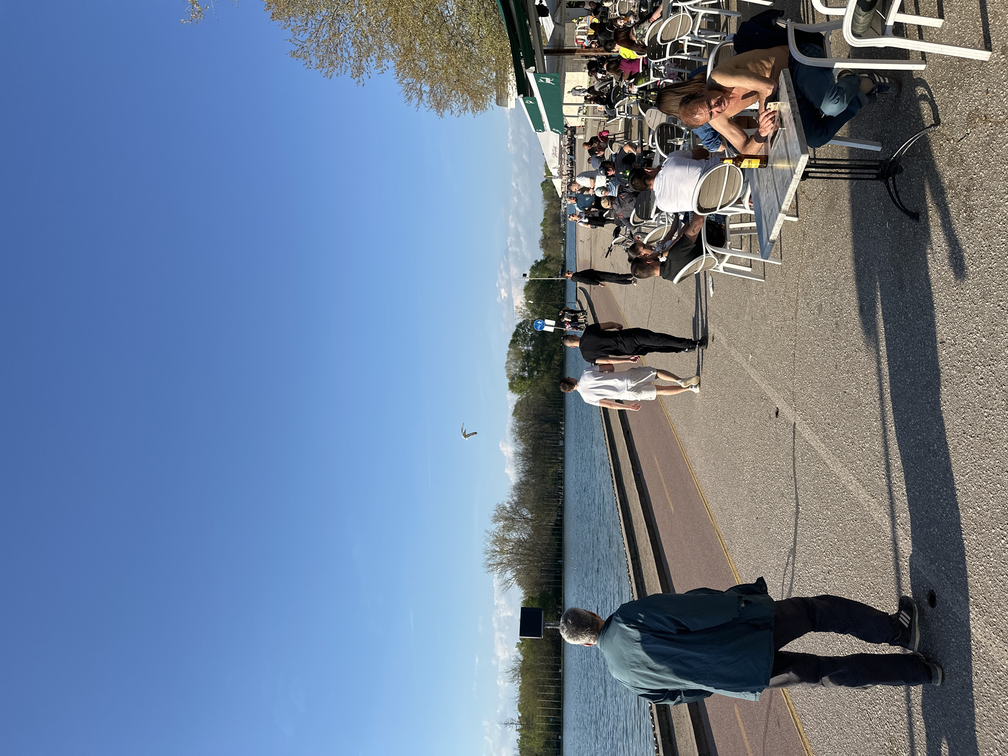 The image depicts an outdoor scene in Plovdiv, Bulgaria, alongside a body of water, likely the Rowing Canal. People are walking along a paved path next to the water, while others are seated at tables under a green awning, possibly a cafe or restaurant. A shirtless man sits at one of the tables. The sky is clear and blue. The sun is shining, casting shadows on the ground. A bird flies in the sky near the water. A man stands near the water. Trees line the water's edge.