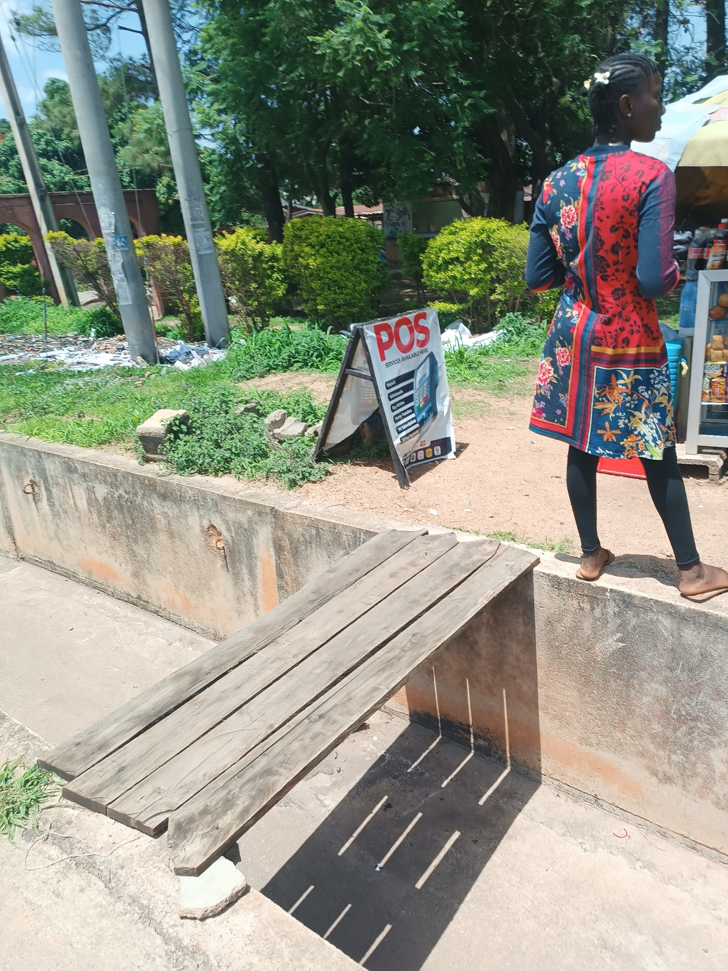 A full shot captures a young woman in a red and blue floral dress with dark leggings and brown sandals, standing on a concrete ledge. Her hair is braided with white beads. She is facing right, away from the camera, and appears to be looking at a display of goods to her right. To her left, a white A-frame sign displays "POS SERVICES AVAILABLE HERE" and lists various financial services like Deposit, Withdrawal, Airtime, and Bills Payment. The background features lush green trees and bushes, several utility poles, and some scattered debris on the ground. In the foreground, a weathered wooden plank structure rests on a concrete barrier, casting sharp shadows from the sunlight. The scene appears to be an outdoor market or roadside stall in the city of Jos, Nigeria, judging by the vegetation and the presence of a POS service sign, common in Nigerian commercial areas.