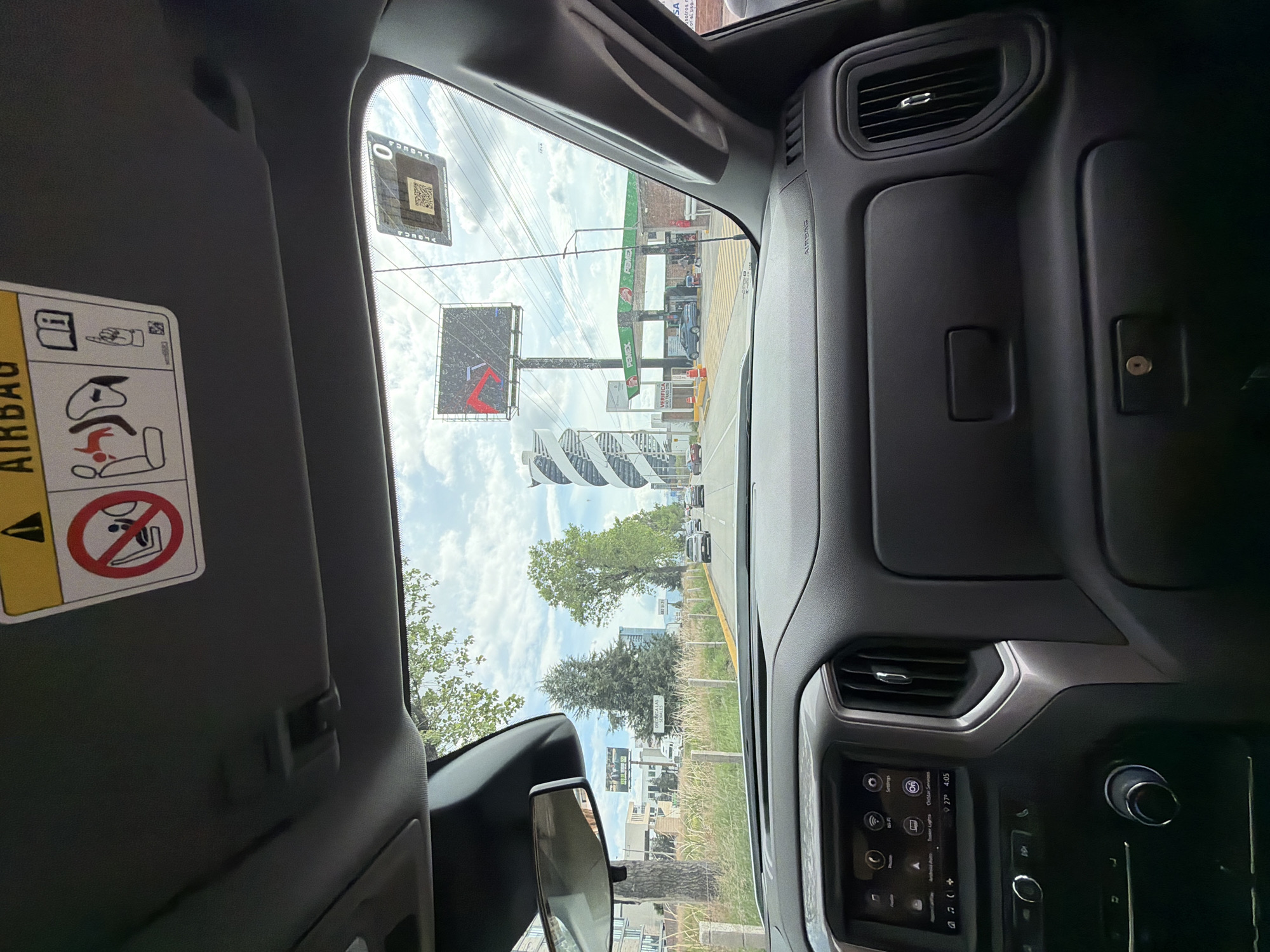 The image captures a view from the interior of a vehicle, looking forward through the windshield. The dashboard is visible in the foreground, featuring a screen displaying infotainment options and various controls. Above the windshield, a sun visor with airbag warnings and safety pictograms is present.
Looking out, the scene depicts a street in San Bernardino Tlaxcalancingo, Mexico. A highway or main road is visible with several cars driving on it. To the right, a Pemex gas station with its distinctive green and red branding is prominent, with vehicles at the pumps and people visible. A large billboard advertising a product or event stands behind the gas station. Further in the background, modern buildings, including a distinctive spiral-shaped structure, are visible alongside trees and overhead power lines, indicative of an urban or semi-urban environment. The sky is partly cloudy.