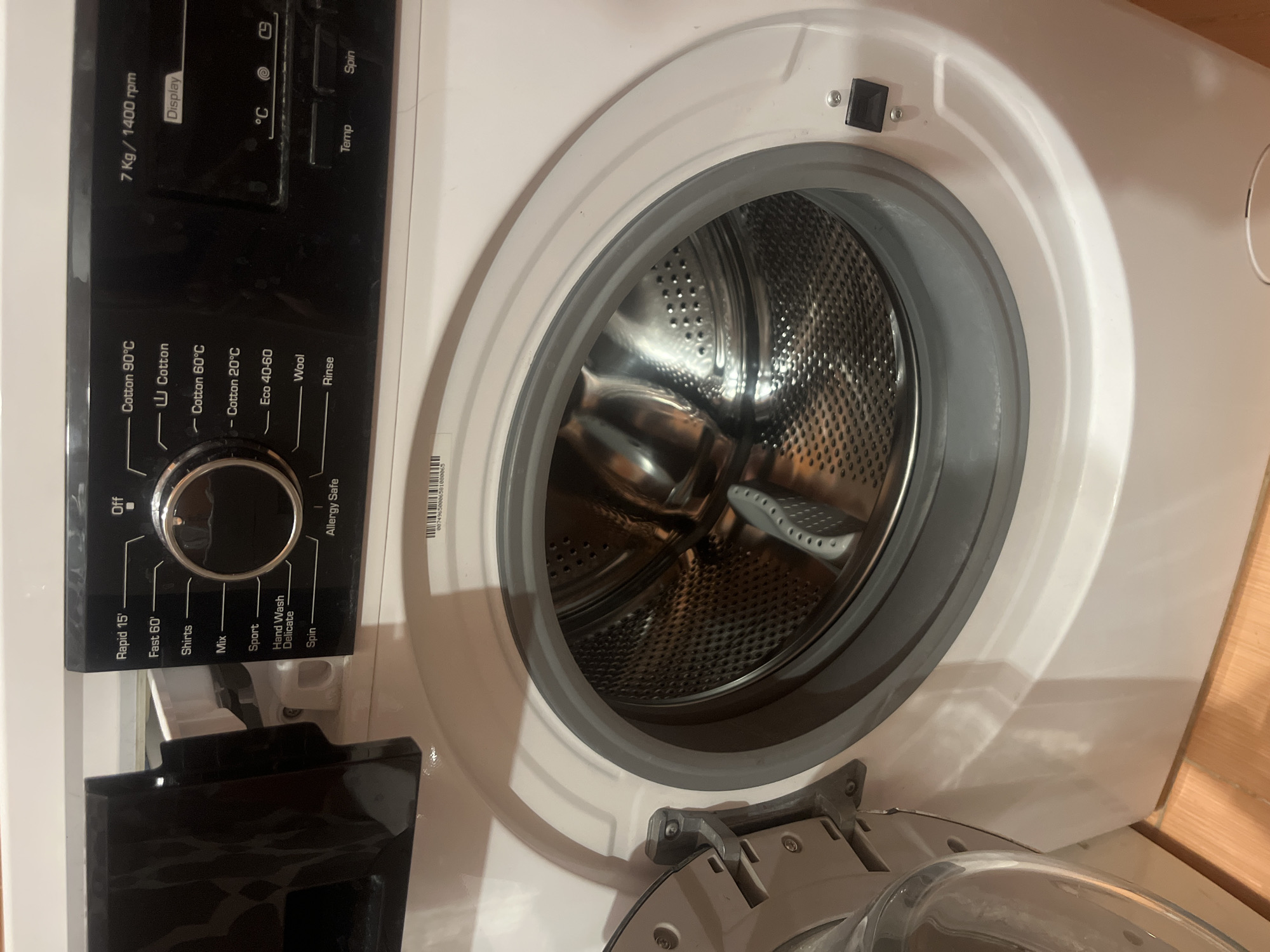 A white washing machine is shown in a close-up view. The washing machine has a black control panel with a dial and various buttons. The door of the washing machine is partially open, revealing the stainless steel drum inside. The washing machine is placed on a light brown wooden floor. The image is well-lit, with no shadows or obstructions. The scene appears to be in a domestic setting.