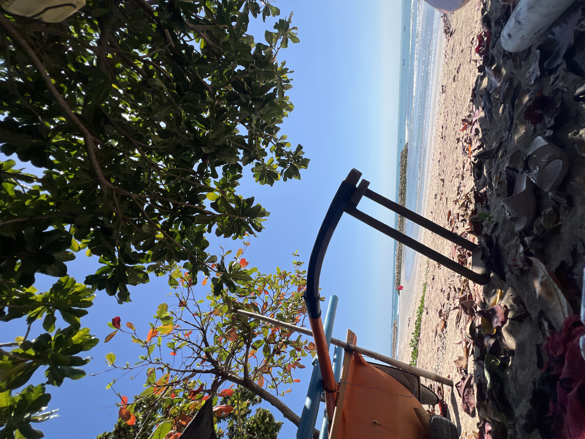 The image depicts a beach scene in Kuta, Indonesia, under a clear blue sky. In the foreground, dry leaves and sand cover the ground, with a pair of sandals and a piece of driftwood visible. Partially obscuring the view is an orange boat on a trailer, supported by wooden legs. The boat has blue and orange pipes attached to it. Beyond the boat, a sandy beach stretches towards the ocean, where gentle waves are breaking. A breakwater made of rocks extends into the sea, and a small red flag is planted on the shore. Several small boats are dotted on the water in the distance. Lush green foliage from trees hangs over the scene from the top of the frame, with some branches also showing leaves that have turned red and orange. The overall impression is of a tranquil beach setting, possibly during a quiet time of day.
