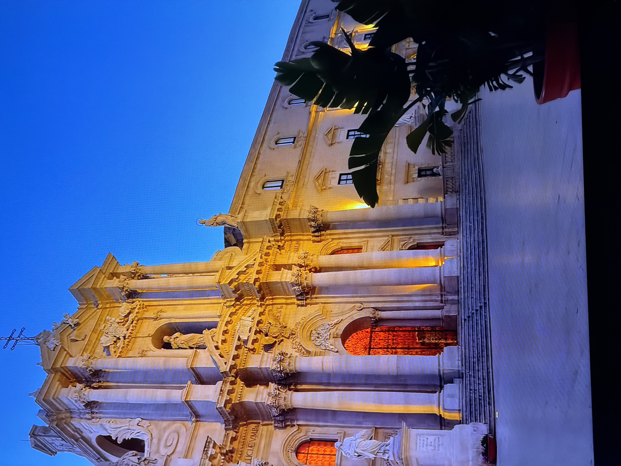 The image depicts the Cathedral of Syracuse, also known as the Duomo di Siracusa, located in the Piazza del Duomo in the city of Syracuse, Sicily, Italy. The cathedral is illuminated by warm, artificial lights, highlighting its Baroque facade. A statue stands to the left of the entrance, and a large potted plant with dark green leaves partially obscures the right side of the building. The sky above is a deep, clear blue, indicating it is either dusk or dawn. The ground in front of the cathedral appears to be a paved plaza, with a set of wide stairs leading up to the entrance. The architectural style of the cathedral is grand and ornate, with columns, sculptures, and intricate detailing.