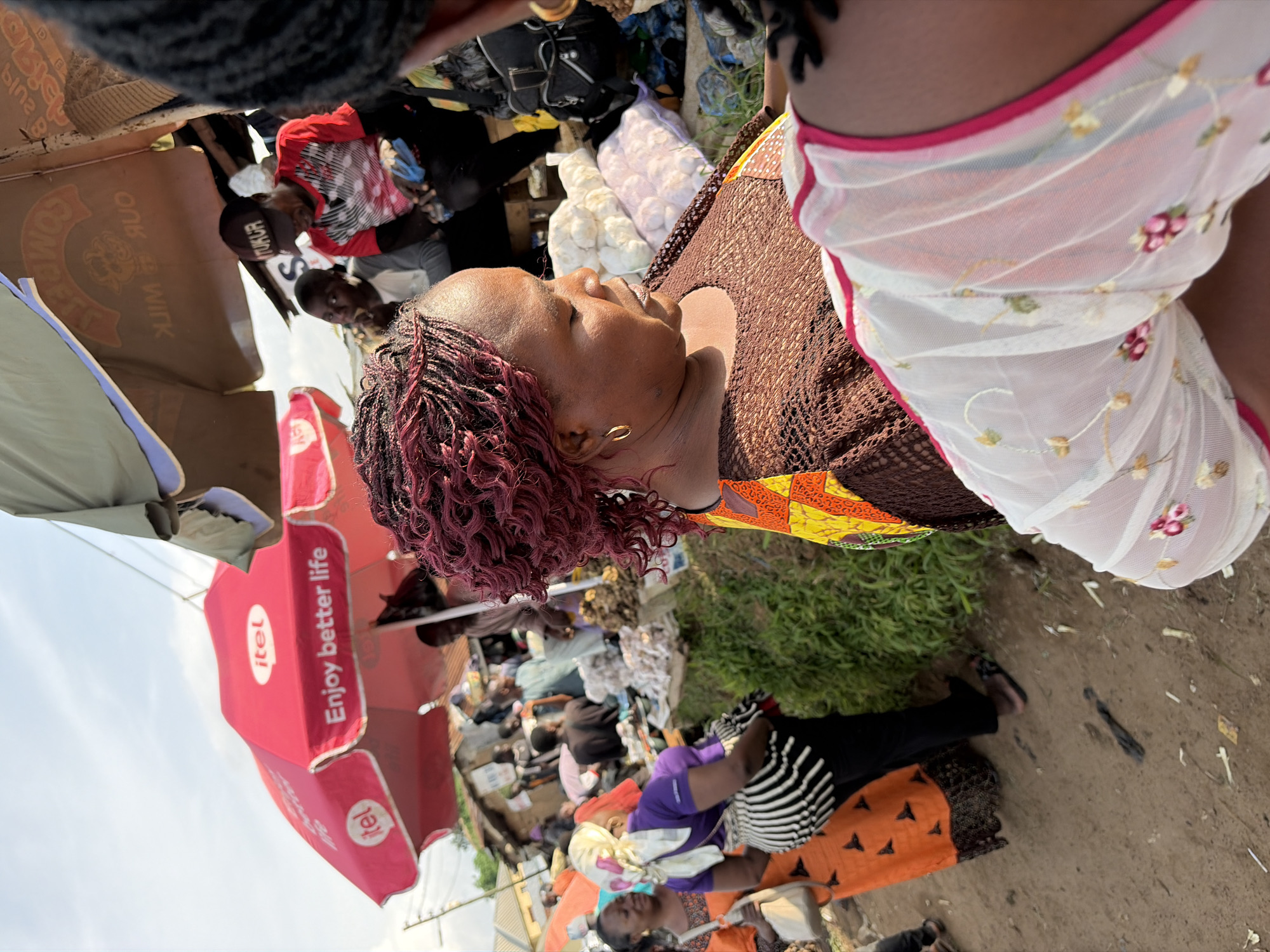 In Gura Top, Nigeria, a bustling market scene unfolds under a bright, overcast sky. In the foreground, the back of a woman with maroon braided hair is visible, wearing a brown, patterned top. To her left, another woman in an orange dress and a white headscarf turns away from the camera. Behind them, various stalls display goods under umbrellas. One prominent red umbrella features the "itel" logo and the slogan "Enjoy better life". Men and women are seen engaged in transactions and moving through the crowded marketplace, with buildings and vegetation forming the backdrop. A stack of white bags, possibly containing produce, sits near the center of the image.