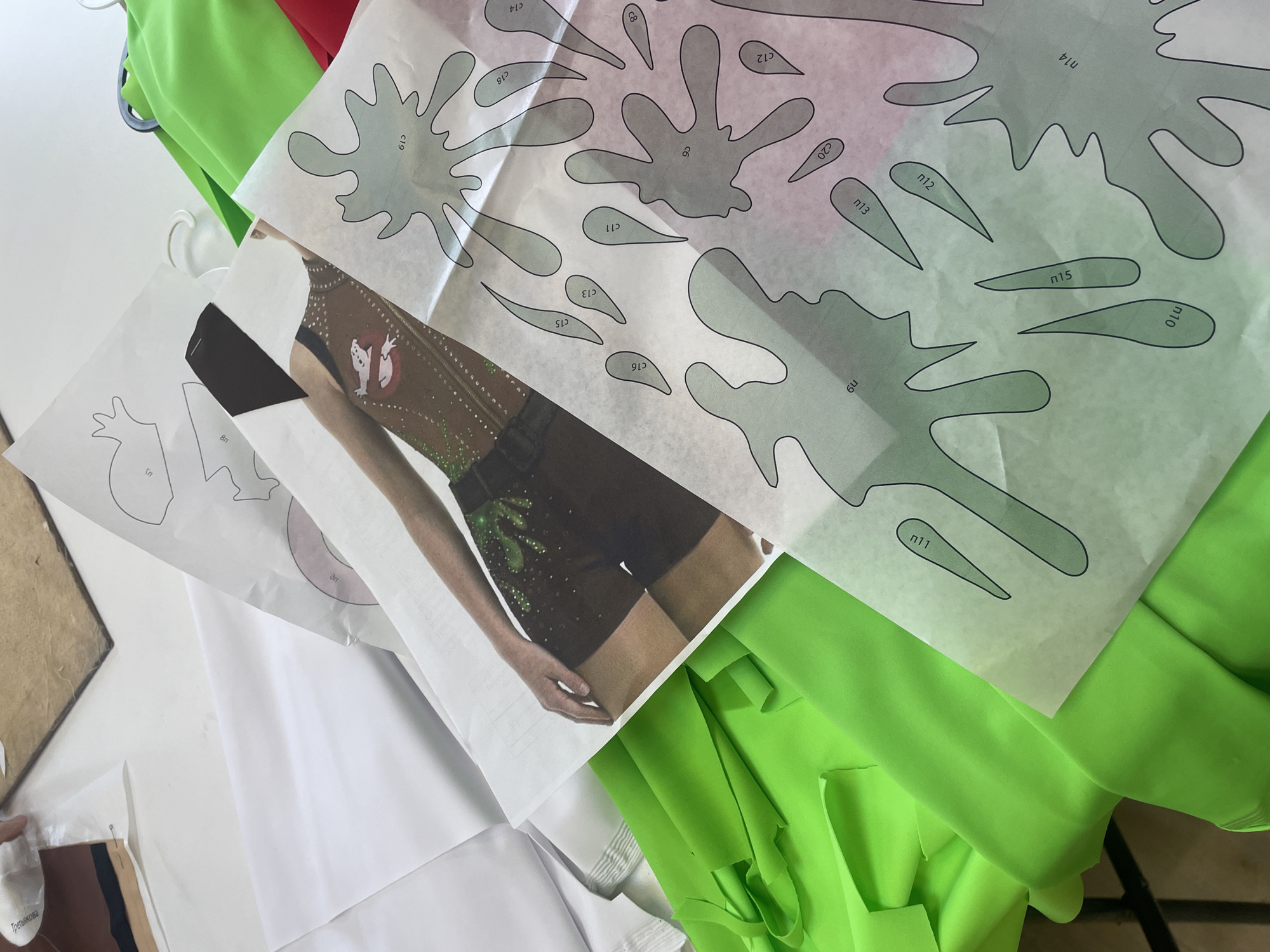 The image depicts a collection of sewing patterns and brightly colored fabrics, specifically neon green and red. The patterns are laid out on top of the fabrics, some of which are white. One pattern shows a figure wearing a costume with a ghost logo, reminiscent of the Ghostbusters franchise. The context suggests a crafting or sewing project is in progress. The environment appears to be an indoor workspace, possibly a studio or home setting. There are no visible clues to suggest a location in Sinyavino, Russia.