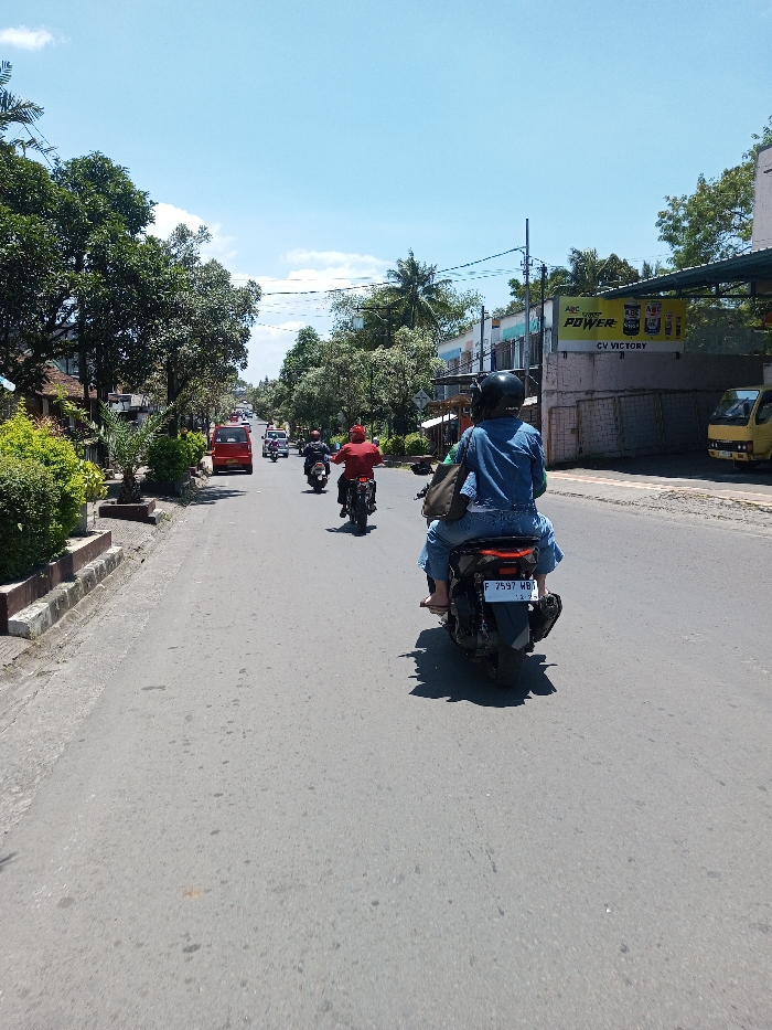 The image captures a daytime street scene in the city of Tagtse Tashidingkha, Bhutan, under a clear blue sky. The viewpoint is from behind a dark-colored scooter moving forward on a grey asphalt road.
In the foreground, a driver wearing a black helmet, blue denim shirt, and blue jeans operates the scooter. A passenger in blue denim, with bare feet, rides directly behind the driver. A dark shoulder bag is visible on the driver. The scooter's rear license plate displays "F 7597 WBA" with "12.23" partially visible below.
Ahead of the foreground scooter, a line of at least three motorcycles proceeds in the same direction, including one rider prominently wearing a red shirt. Further down the road on the left, a red van or SUV is visible, followed by a lighter-colored car.
The roadside on the left features a concrete curb and raised planters with green bushes and trees. The right side of the road is lined with buildings, including one with a large yellow and black sign displaying "ABC POWER," "ABC OIL," and "CV VICTORY." A yellow utility truck is parked or stopped further down the right side of the road. Utility poles with overhead wires are visible along the right side, and abundant green trees, including palm trees, line the street. The road surface appears dry and clear.