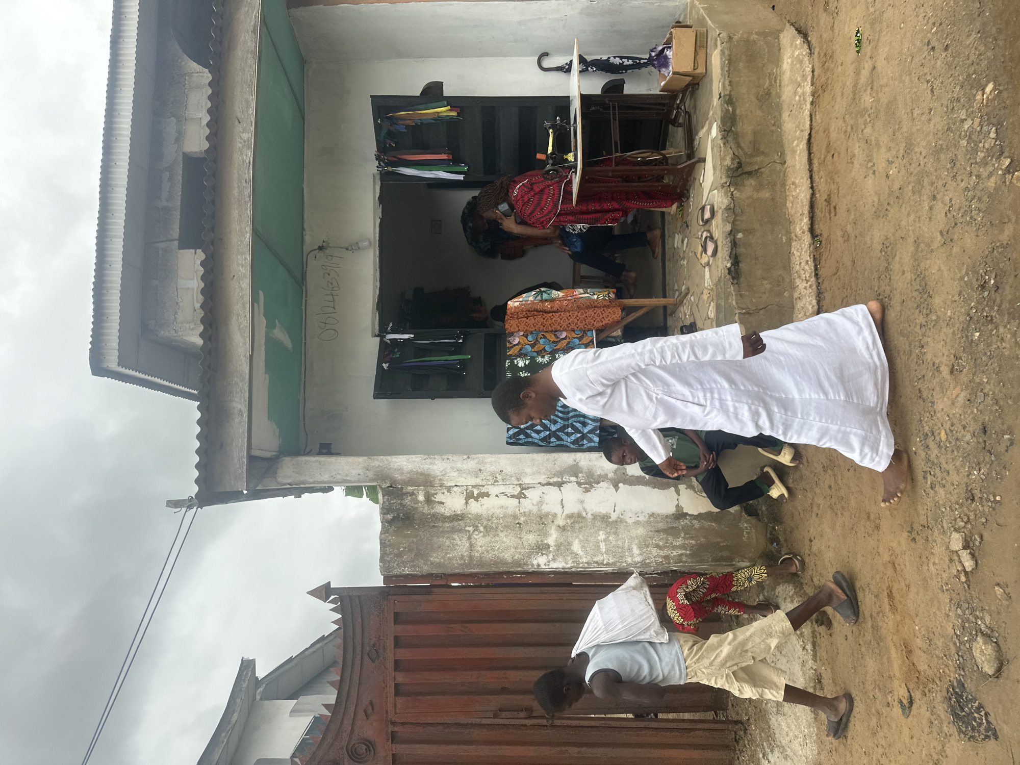 The image depicts an outdoor scene in Ajara, Nigeria, with a focus on a small tailor shop. The shop appears to be a modest building with a corrugated metal roof and a light-colored facade. Inside, a woman in a red and black patterned dress is seated at a sewing machine, actively engaged in tailoring. Various fabrics, some folded and stacked on shelves, and a selection of colorful umbrellas are visible within the shop.
Several people are present in front of and around the shop. Two individuals, both appearing to be men, are standing near the entrance. One, wearing a white robe, is facing away from the camera and looking down, possibly at the ground or at someone. Another person, partially visible in a green shirt, is seated nearby. Further down, a young boy in a light blue shirt and tan shorts is lying on a metal gate, appearing to be resting or waiting. Another person in a red and yellow patterned skirt and white top is also visible near the shop.
The ground is uneven and appears to be dirt or sand, with some scattered gravel. The weather seems overcast, suggesting a cloudy or possibly rainy day. The overall atmosphere is casual and suggests everyday life and commerce in a Nigerian community. There is some visible text on the wall of the shop: "08124623919". The image is oriented vertically.