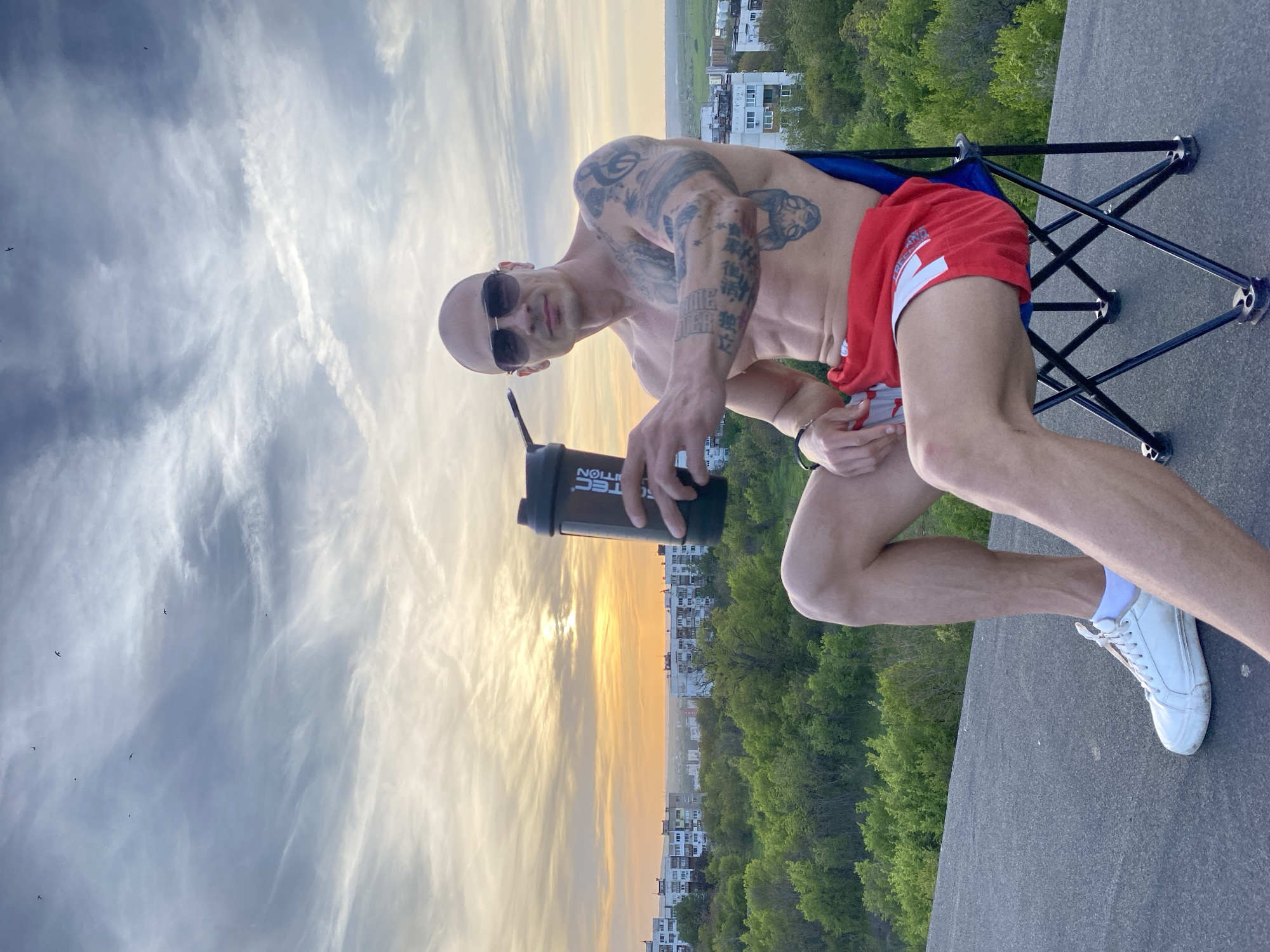 A bald, muscular man with extensive tattoos on his upper body poses on a rooftop during what appears to be sunset, overlooking a cityscape of Pleven, Bulgaria. He is shirtless, wearing sunglasses, red athletic shorts with a white logo, and white sneakers. The man is seated on a blue and black portable folding chair and holds a black shaker bottle with the partially visible text "SCITEC NUTRITION" in his right hand, looking directly at the camera.
His tattoos include various designs on his arms and shoulders, with visible Asian script on his right arm and chest, and the words "GAME OVER" on his left arm. The background features a dramatic sky with a warm, golden glow from the setting sun, fading into cooler blue tones with scattered clouds and faint contrails. Several small birds are visible in the sky. Below the horizon, a sprawling urban landscape of residential buildings and abundant green trees stretches into the distance, providing a scenic backdrop to the relaxed rooftop scene.